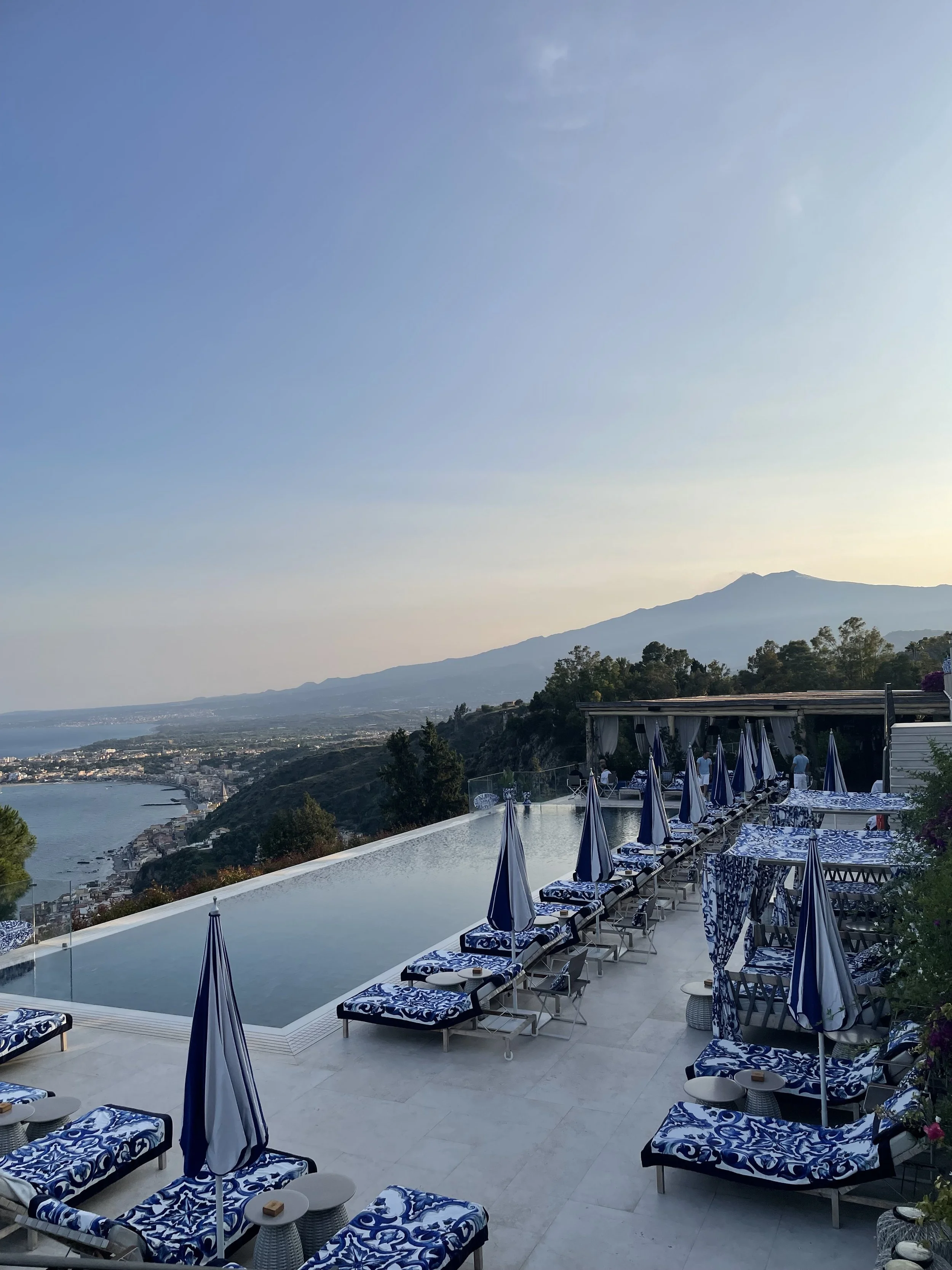 Luxury outdoor pool area with blue and white patterned lounge chairs and umbrellas, overlooking a coastal city and mountains in the distance during sunset.