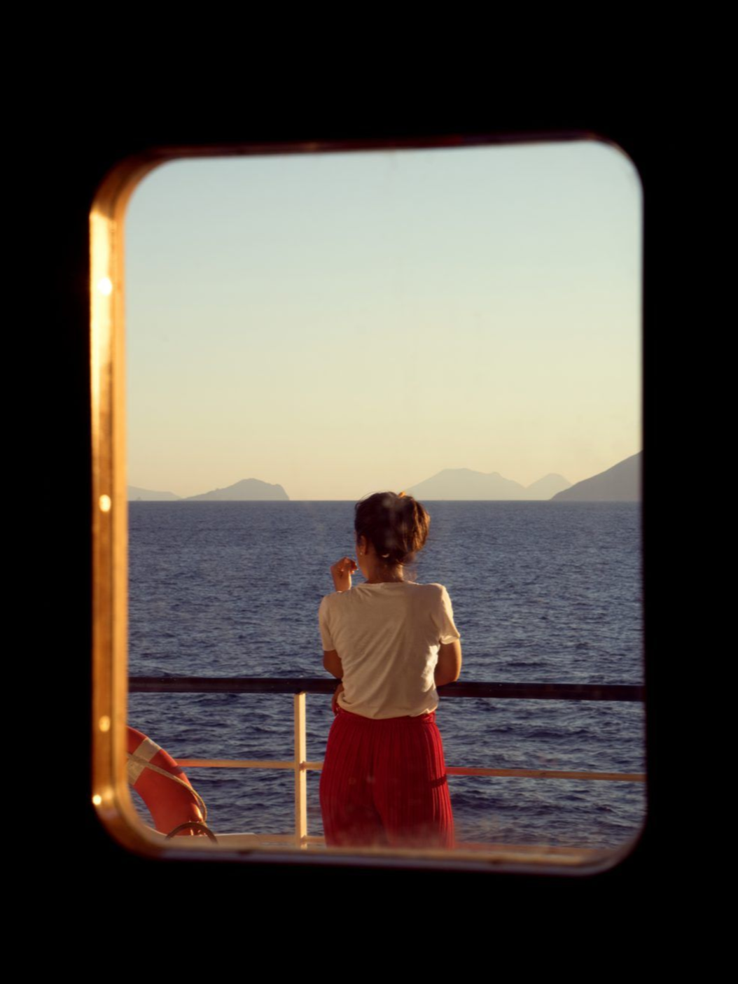 A woman standing on a boat looking out at the sea and distant islands during sunset, viewed through a window.
