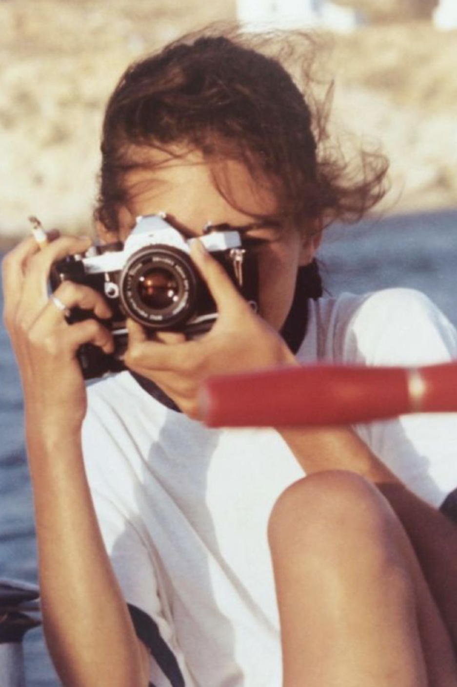 A woman with dark hair holding a camera up to her face, taking a photo outdoors near water.