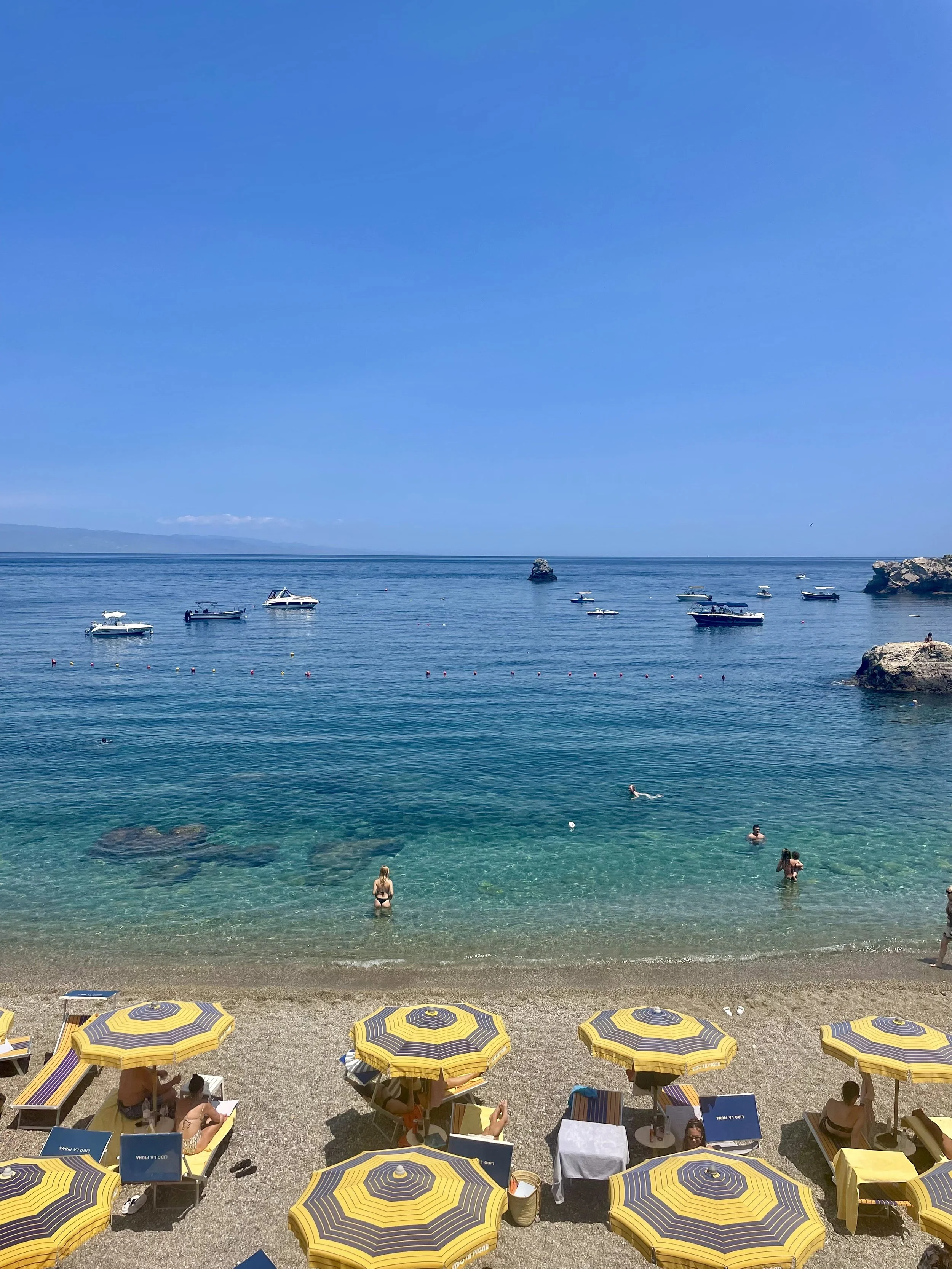 Beach scene with yellow and purple striped umbrellas, people swimming and relaxing near the shore, boats anchored beyond, rock formations, and a clear blue sky.