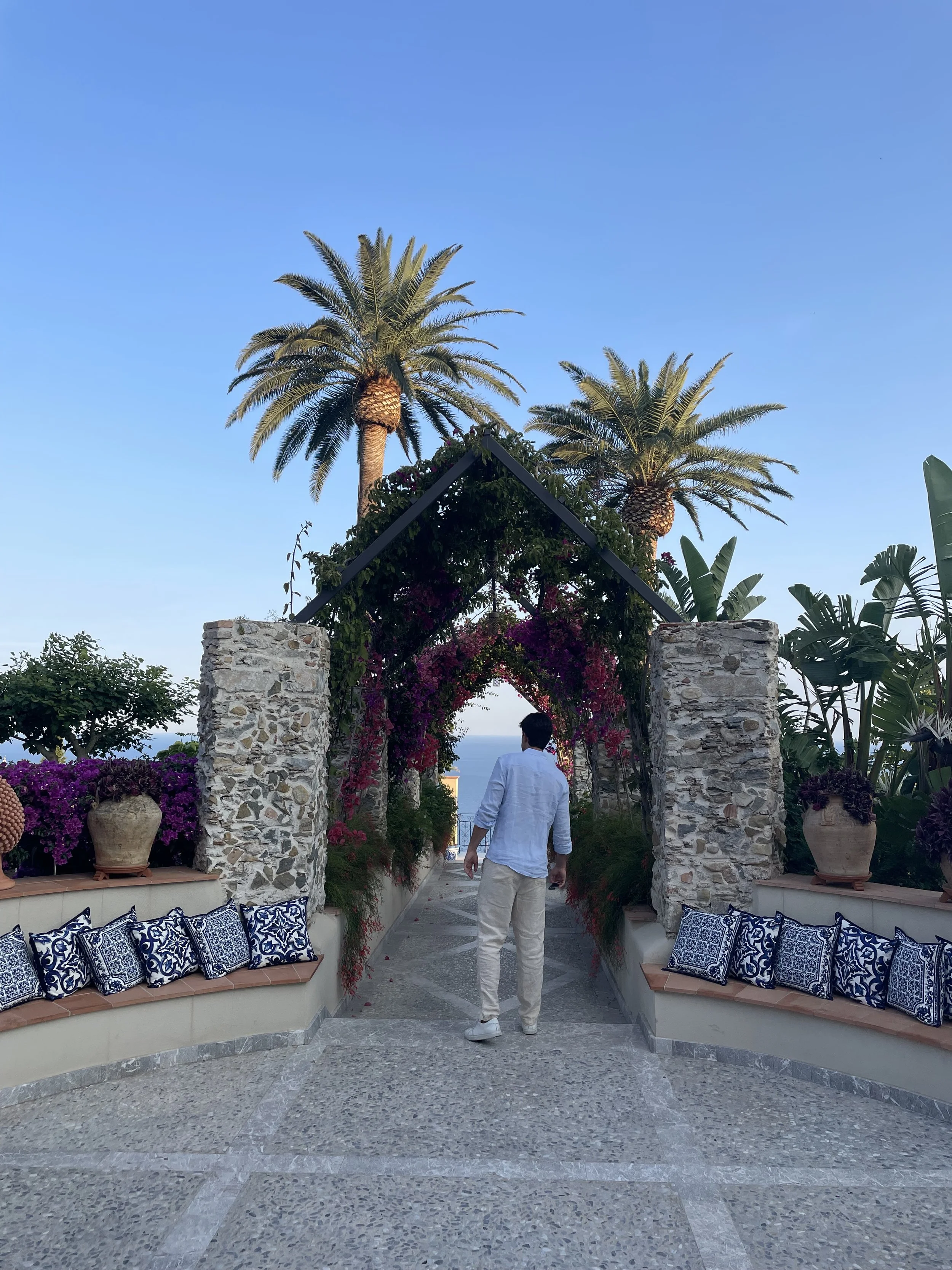 A man dressed in white walking through a garden archway with palm trees and potted plants, overlooking the ocean at sunset.