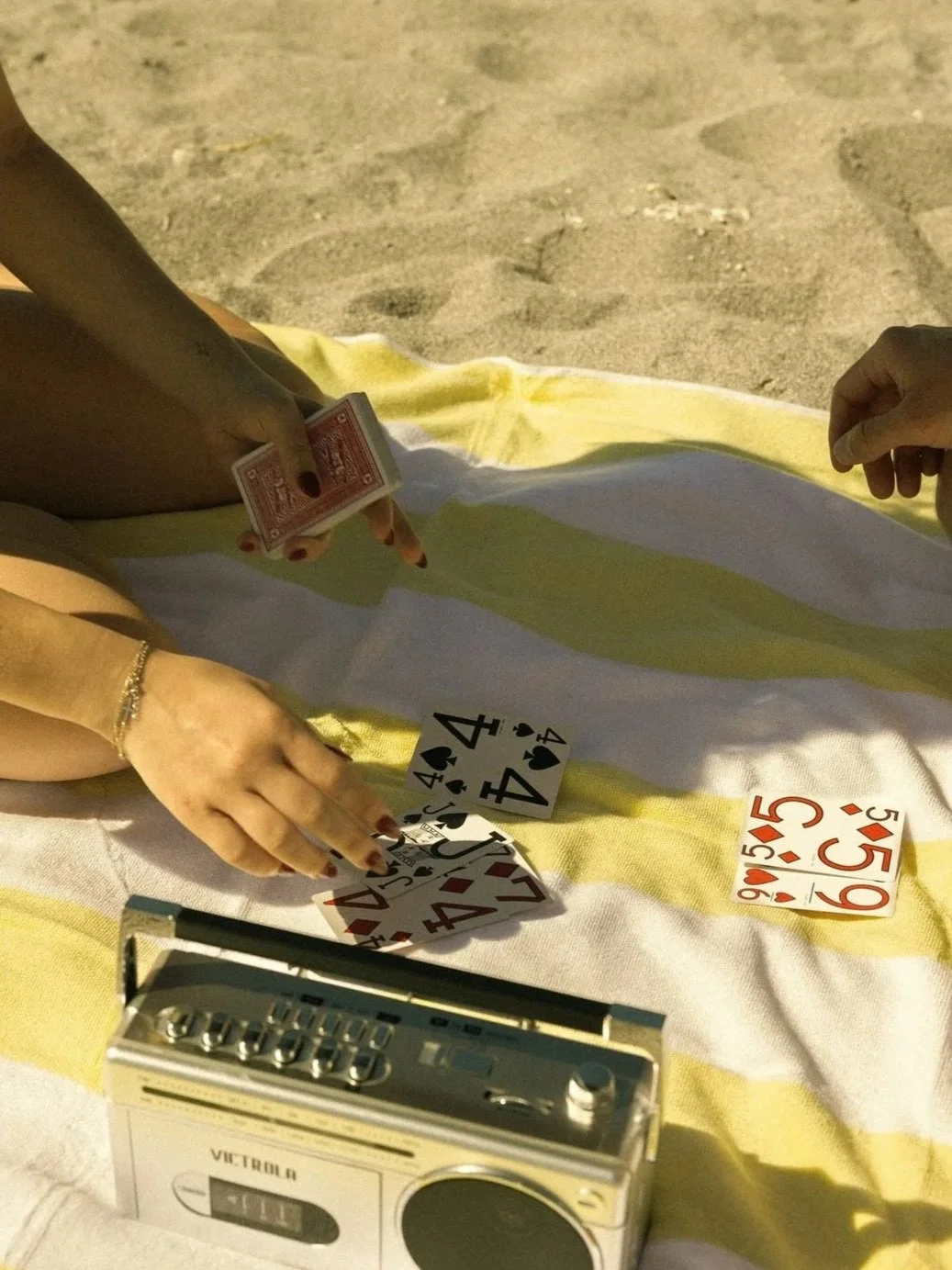 Two people are playing a game of cards on a beach towel. There are scattered playing cards, including fours, fives, and jacks. One person is holding a card, and an old radio can be seen in the foreground.