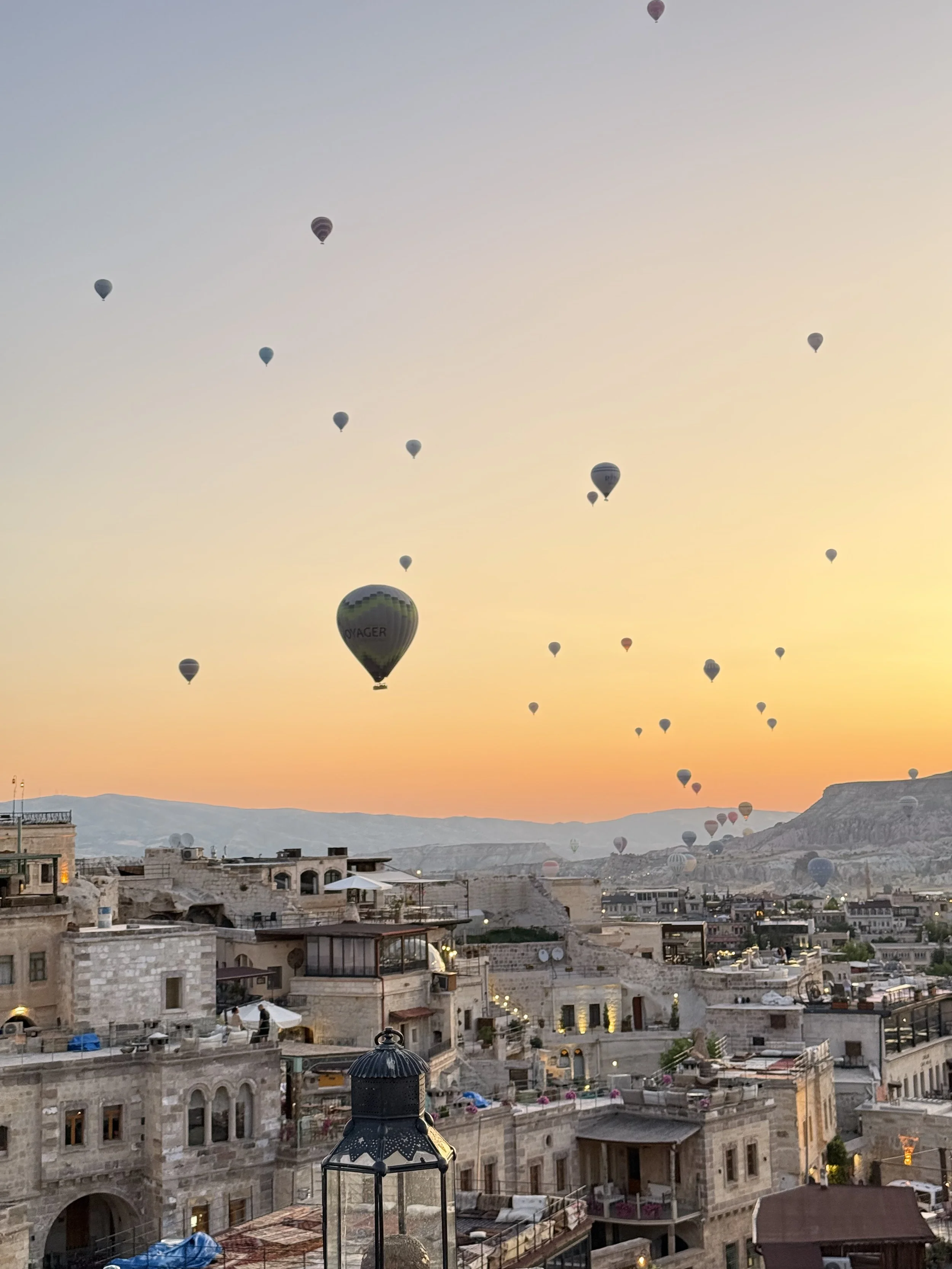 Hot air balloons floating in the sky above a desert city at sunset, with traditional stone buildings and rolling hills in the background.
