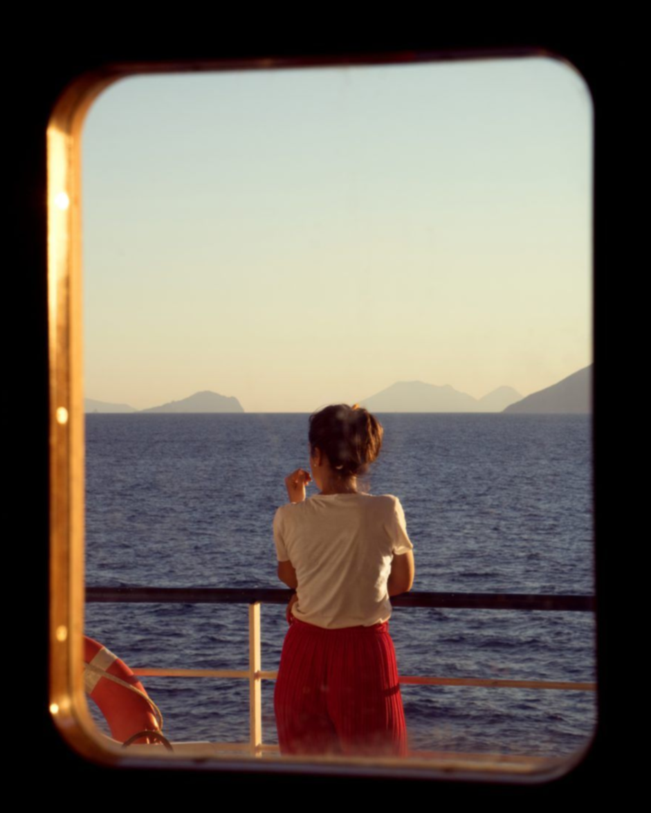 A woman with dark hair in a bun is standing on a boat, looking at the sea and distant mountains through a porthole at sunset, wearing a white t-shirt and red pants.