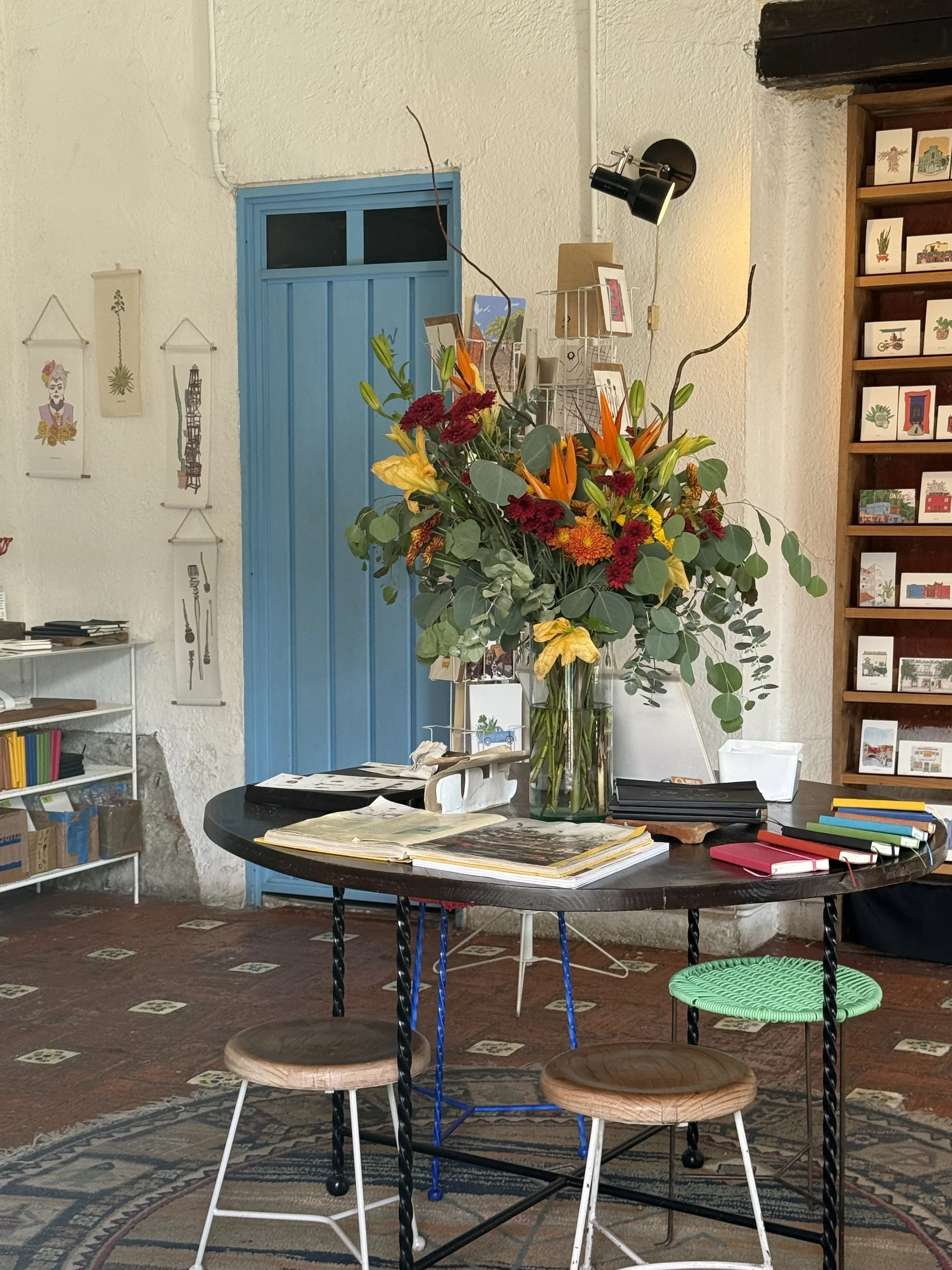 A large bouquet of colorful flowers on a round table indoors, with a blue door in the background and artwork on the walls.