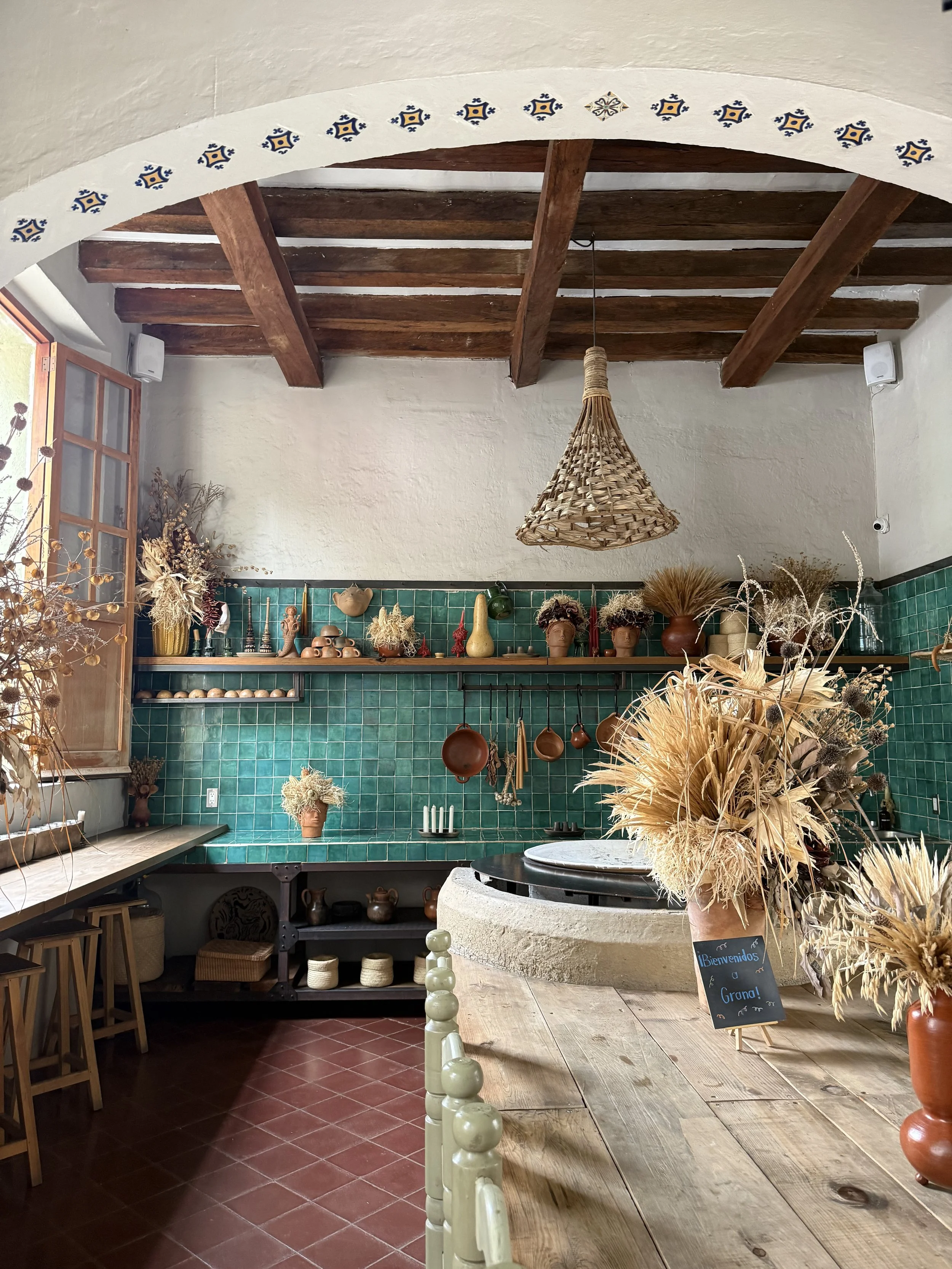 A rustic kitchen with wooden ceiling beams, green tiled walls, and a wooden table decorated with dried flowers and modern pottery, featuring hanging copper pots and a woven pendant light.