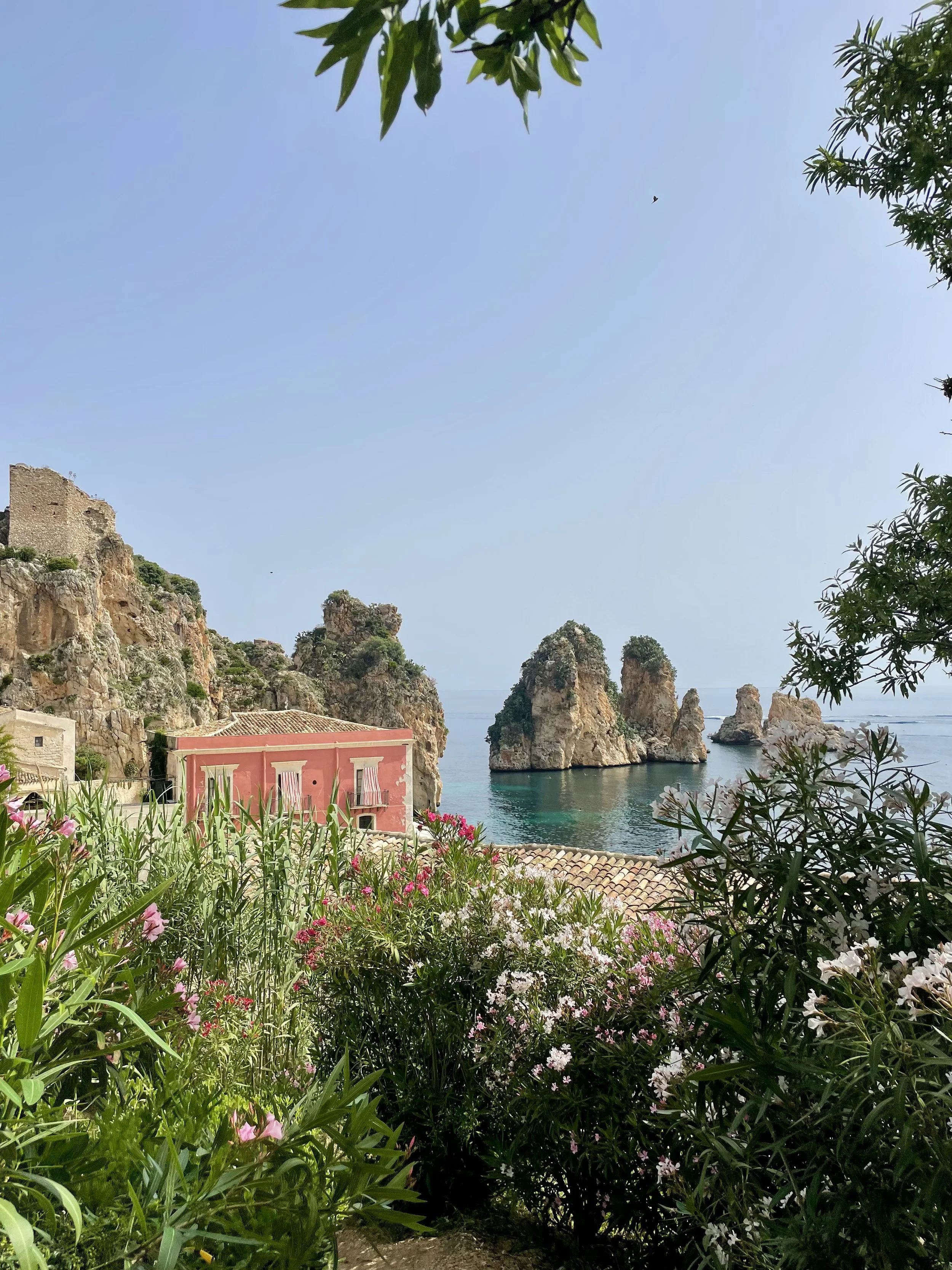 View of a coastal landscape with rocky cliffs and several small island formations in the ocean. Pink and white flowers and green foliage frame the foreground. A pink building with small windows and a tiled roof is visible below the rocks, under a clear blue sky.