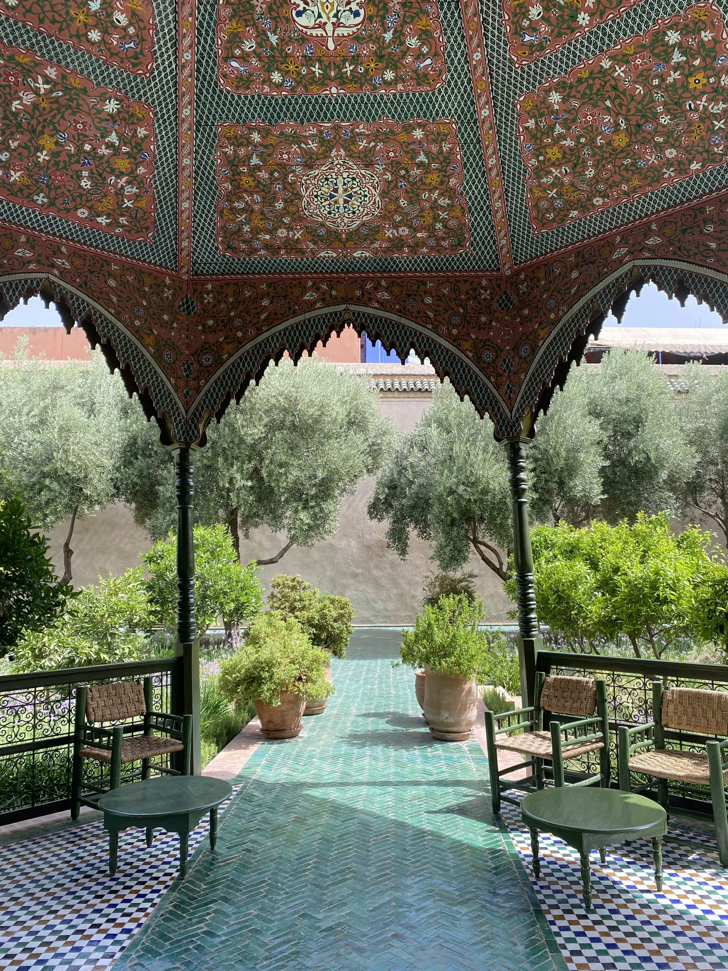 A porch with a decorative Persian-style canopy overlooking a lush garden with olive trees and green plants, and patterned tiles on the floor.