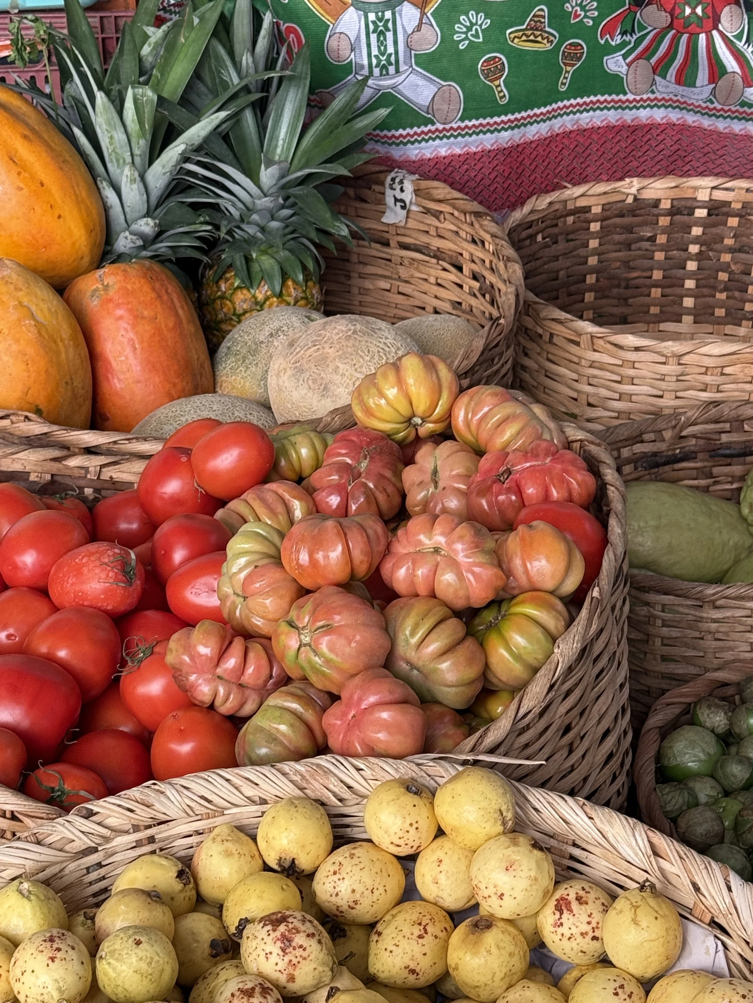 Basket of heirloom tomatoes, with pineapples and cantaloupes in the background, along with other fruits in woven baskets, and a festive tablecloth.