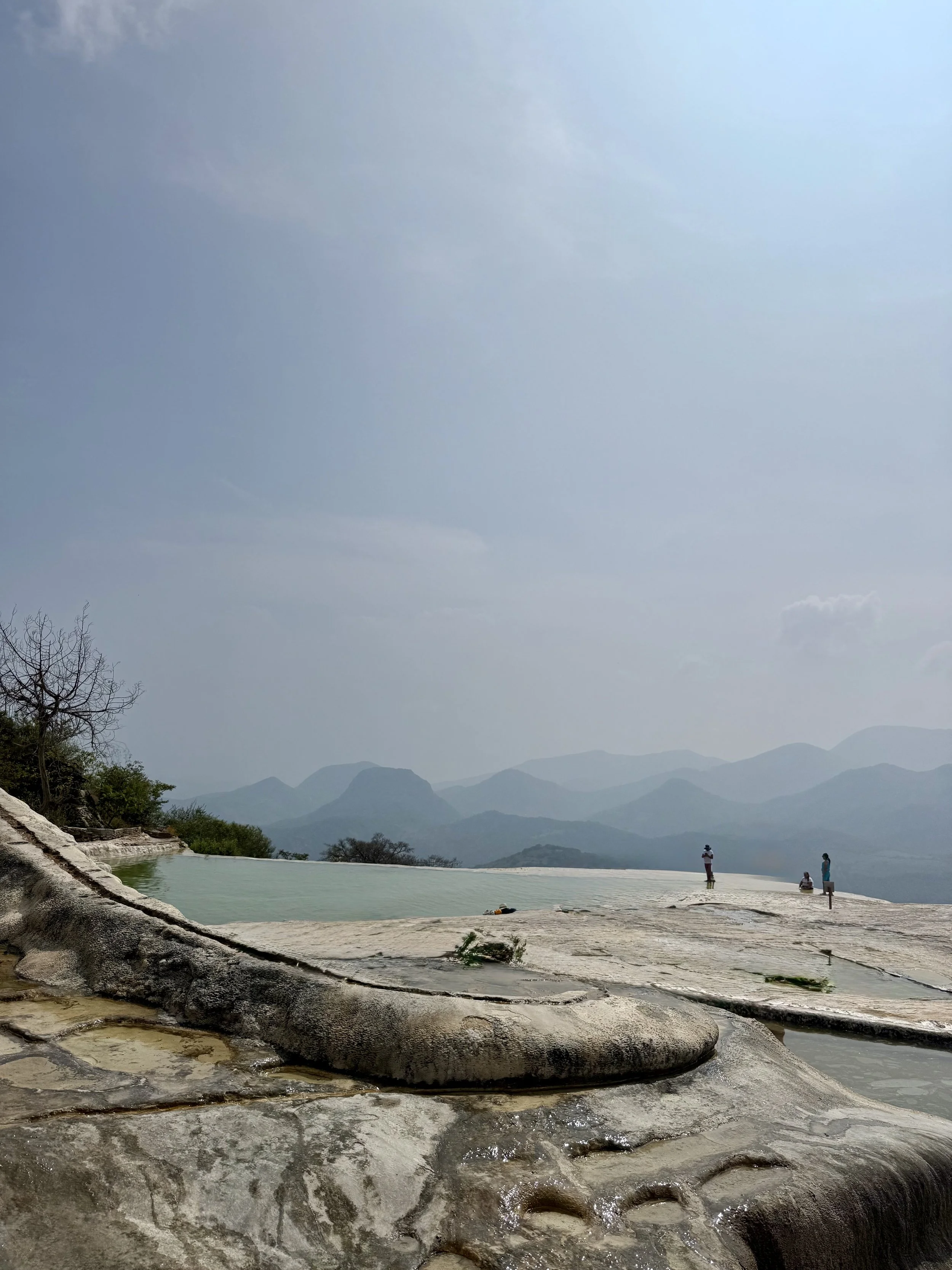 People standing near a mountain-fed water source with distant mountains and hazy sky in the background.