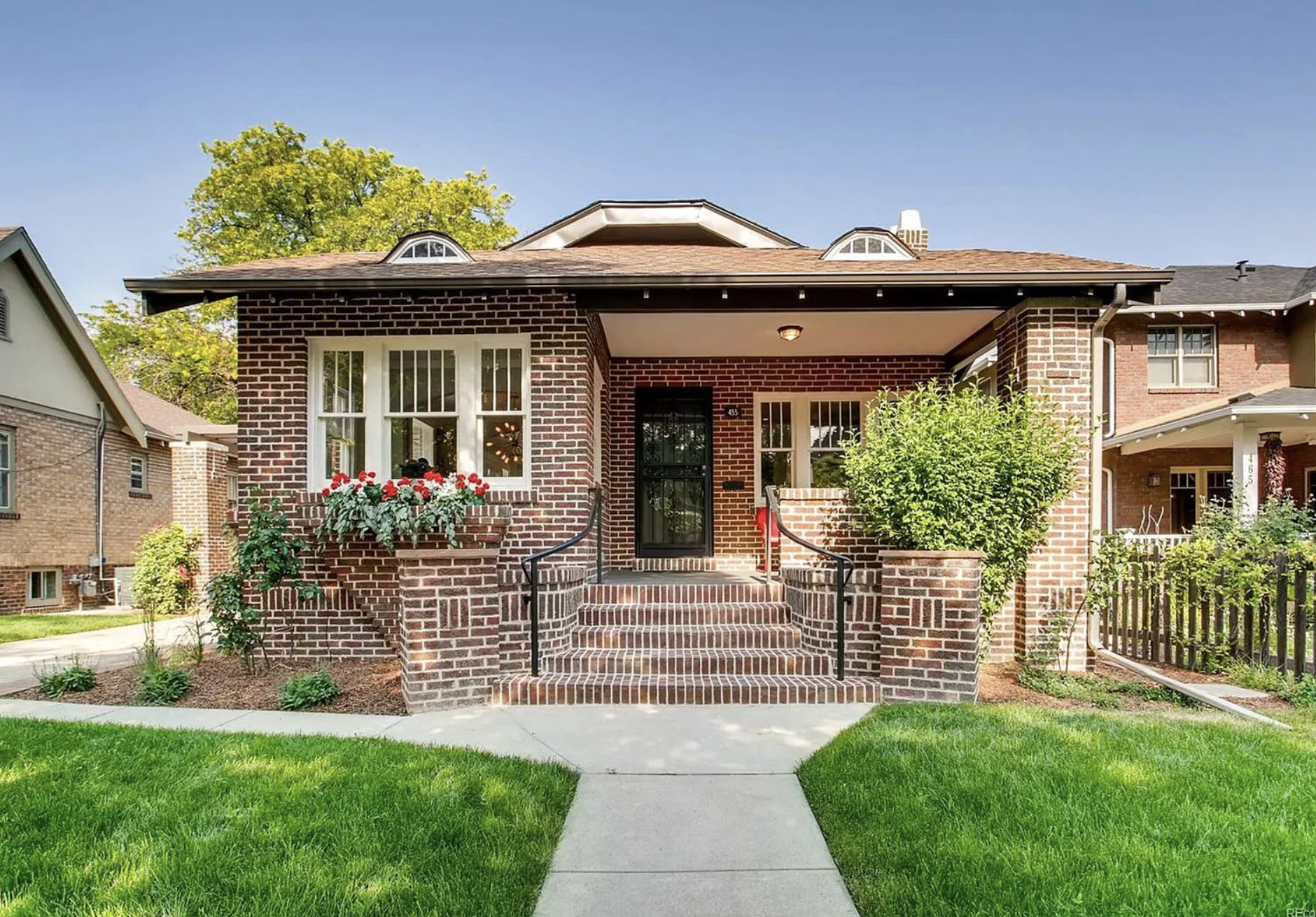 Front view of a brick house with a porch, stairs, and a black door. There are window flower boxes with red and white flowers and green plants. A concrete walkway leads to the front steps, with well-manicured grass on either side.