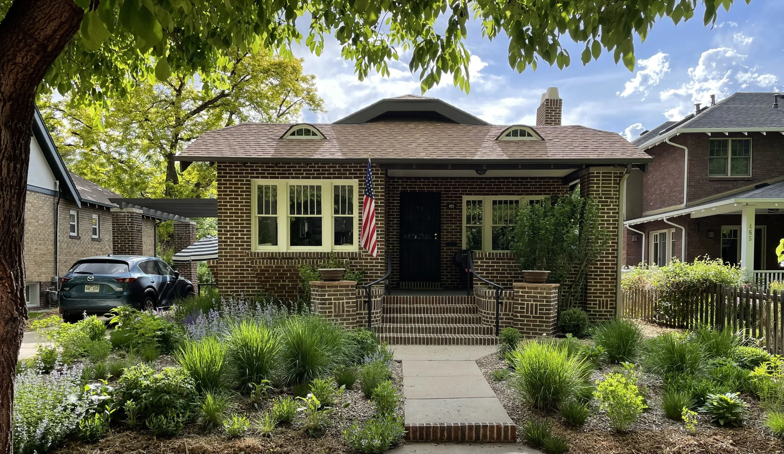 Brick house with steps leading to front door, American flag on porch, lush garden in front, neighboring houses, trees, and blue sky with clouds.