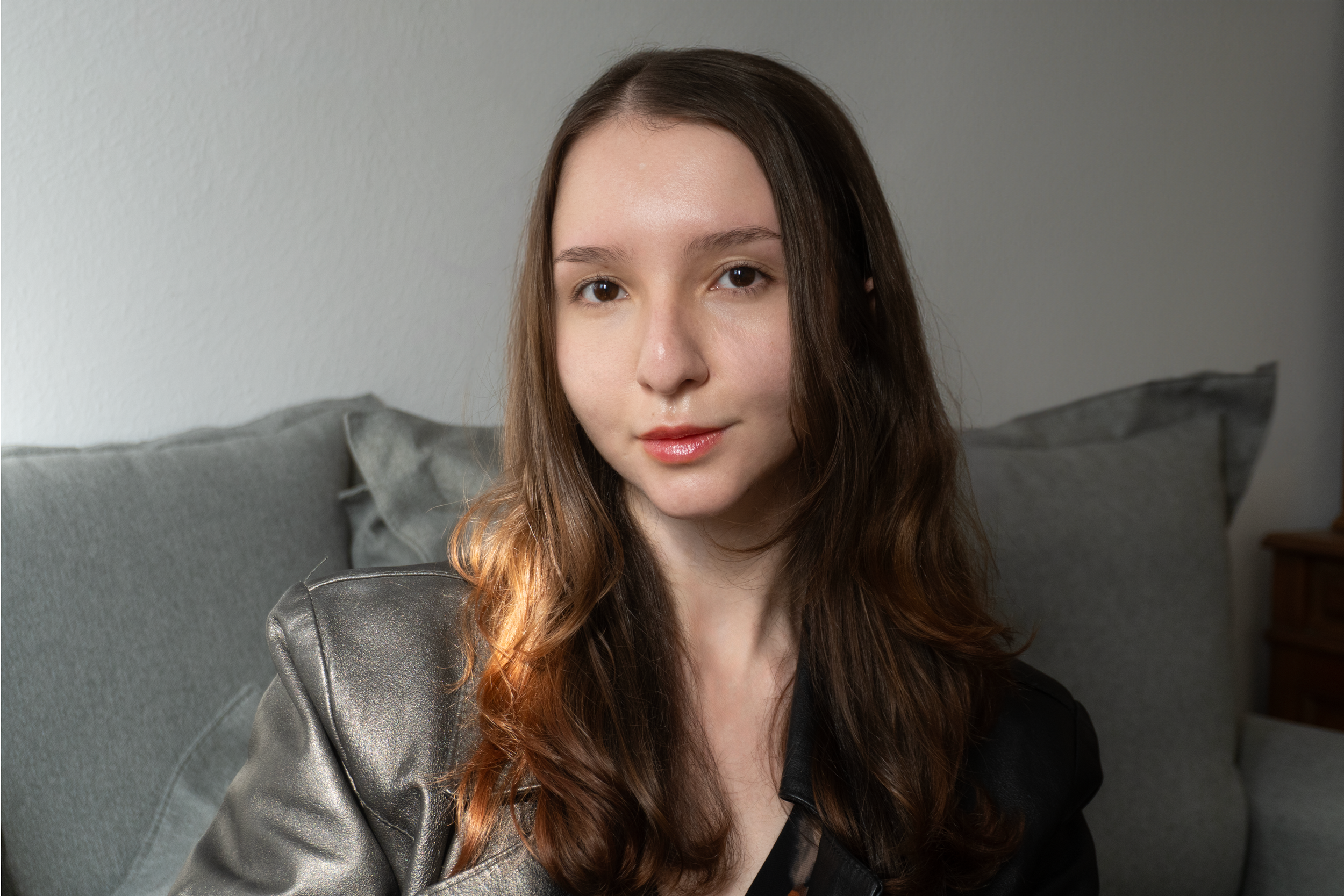 Retrato de uma jovem mulher com cabelo castanho e cabelo ondulado, vestindo jaqueta de couro cinza, sentada no sofá cinza, olhando para a câmera com expressão neutra.
