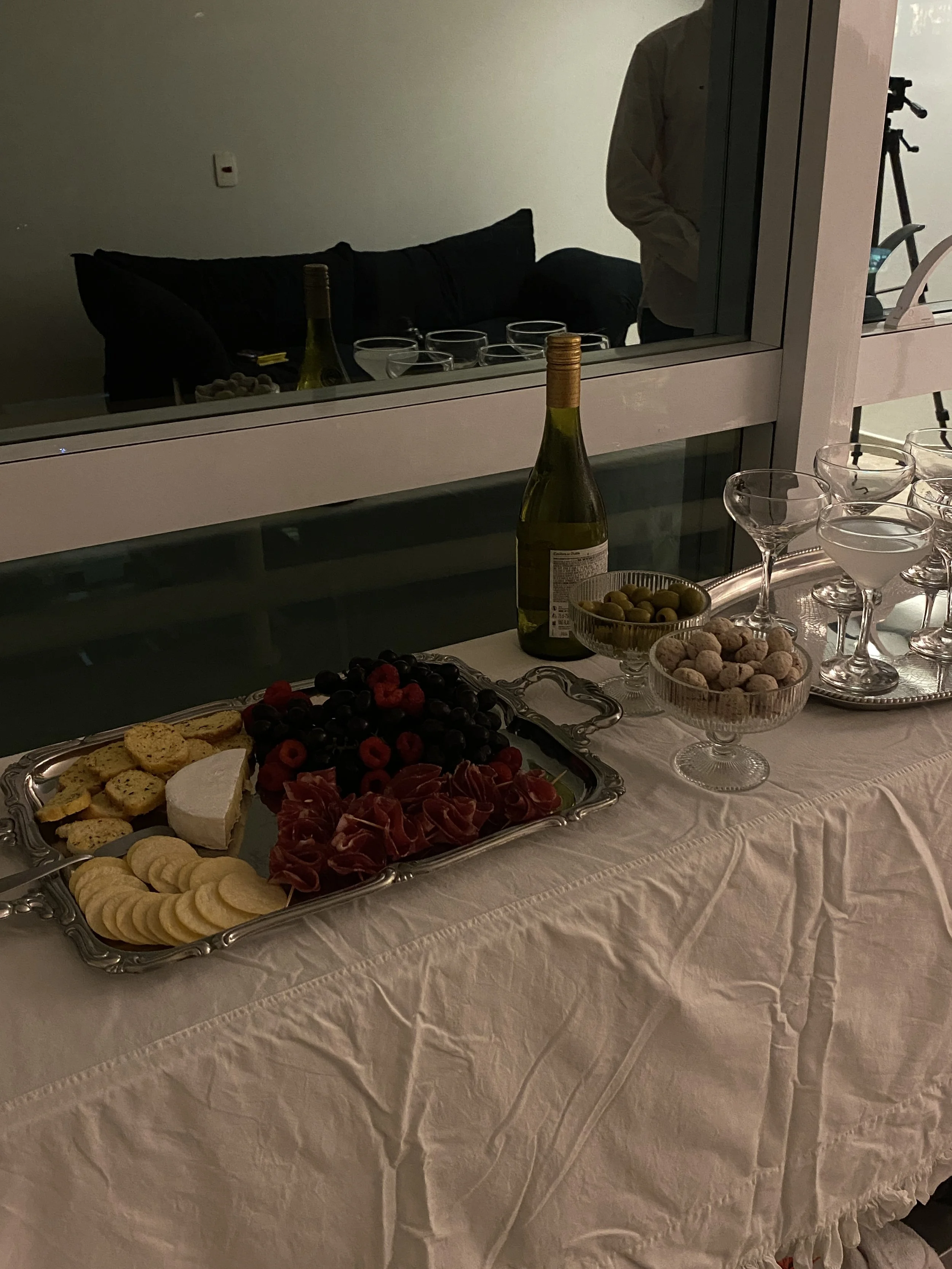 Mesa de festa com bandeja de queijos, presunto, uvas, bolinhas de pão, vinho e copos em ambiente interno com iluminação fraca, refletida na janela.