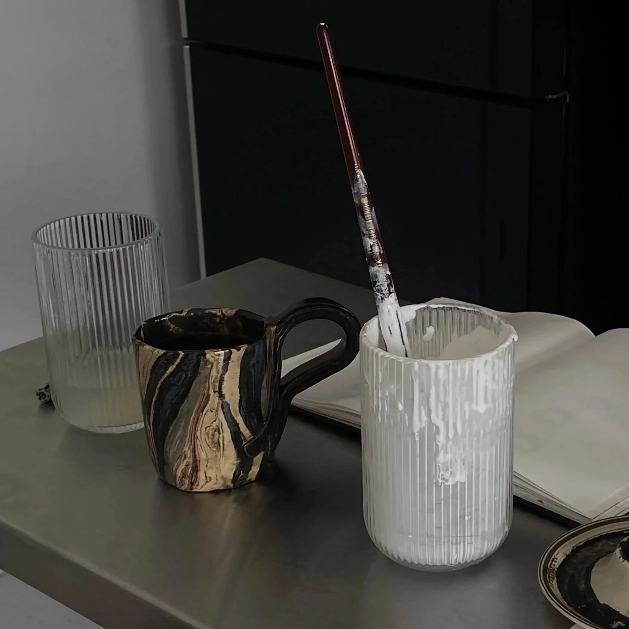 Estes objetos incluem uma caneca de vidro com uma escova de pintura dentro, uma caneca de cerâmica com um padrão marmorizado em cores pretas, brancas e douradas, um copo de vidro cilíndrico vazio com linhas verticais, uma pia de metal com tampa e uma maçaneta, uma tigela de cerâmica com padrão similar ao da caneca marmorizada, uma tigela rasa de vidro na parte inferior, uma tigela de vidro preta e dourada, um carretel de fita de vídeo, e um caderno aberto sobre a mesa.