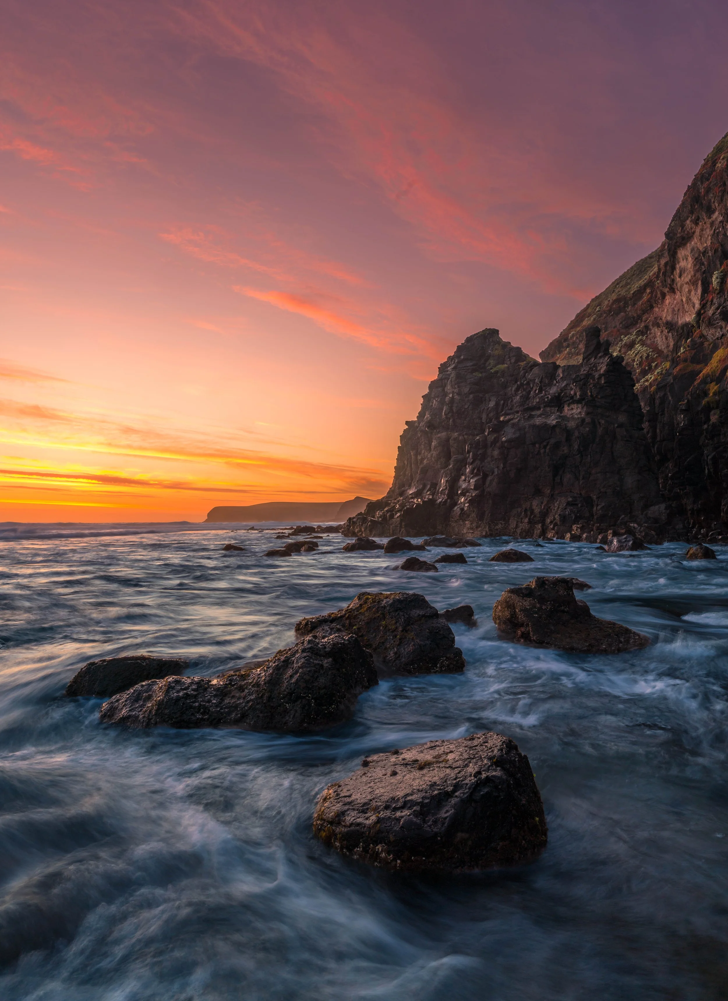 Sunset at Cairns Bay on the Mornington Peninsula, with rugged coastal cliffs, pink and orange clouds, and waves flowing around dark rocks in the foreground.