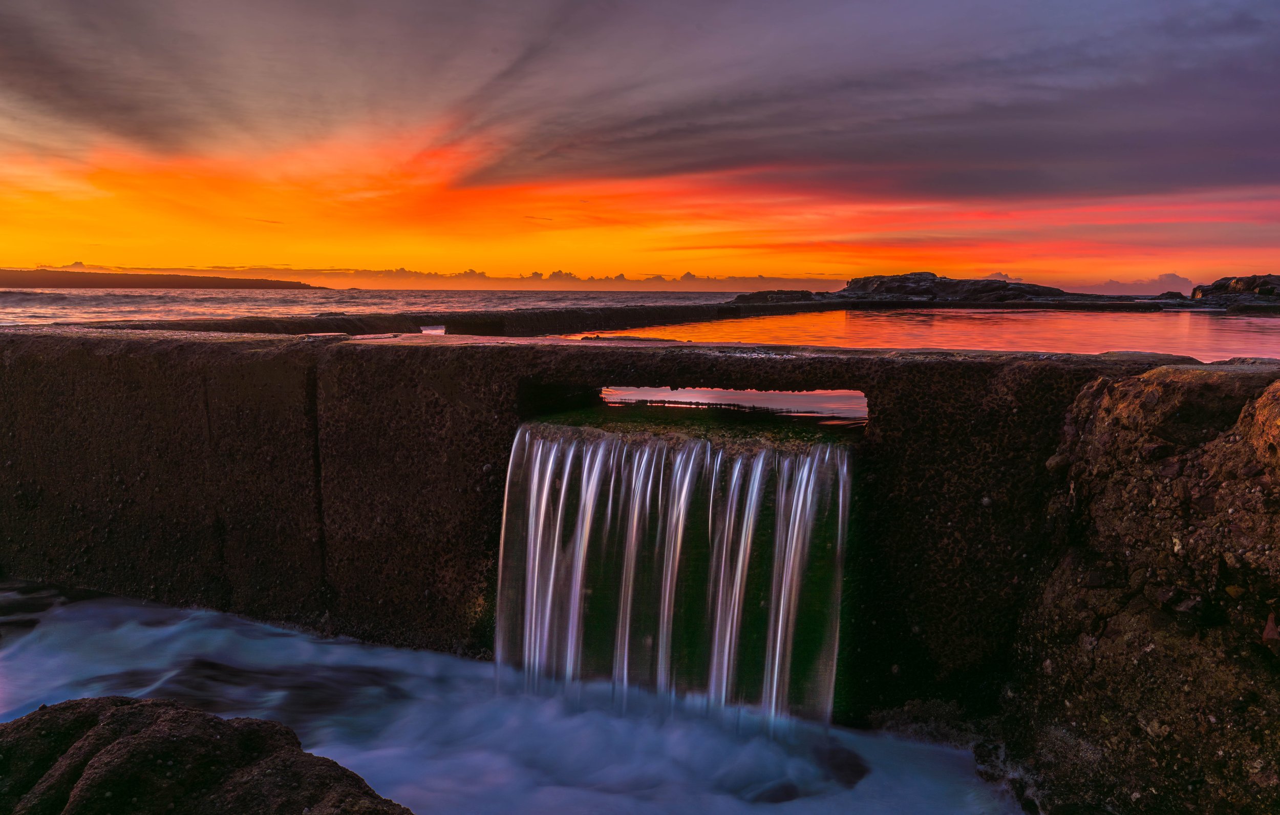 Sunrise at Aslings Beach in Eden, NSW, with a vivid orange and pink sky above the ocean and seawater cascading over a rocky ledge in the foreground.