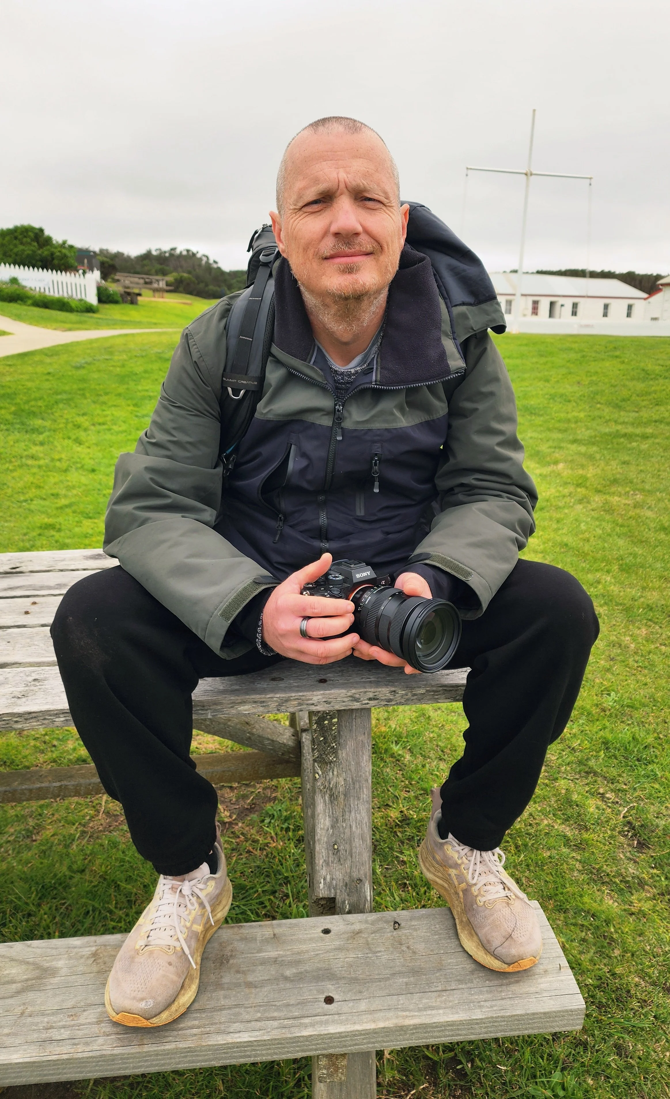 Man sitting on a wooden bench outdoors, holding a Sony camera, wearing a gray jacket, black pants, and tan sneakers, with a backpack on his shoulder and a cloudy sky in the background.