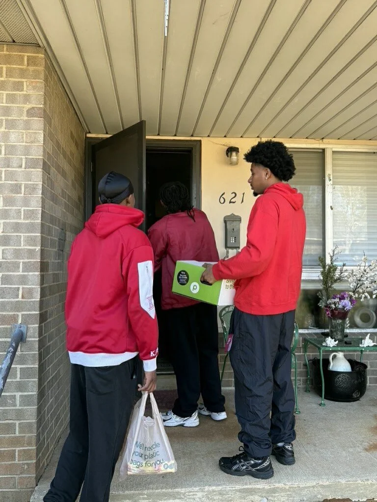 Two people are delivering a box of food to a woman at her front door while another person waits nearby with a bag of groceries.