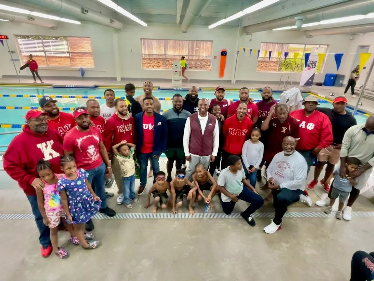 Group of people, including children and adults, posing by an indoor swimming pool for a photo. Some individuals are wearing red and maroon clothing with Greek letters, possibly representing a sorority or fraternity. The background shows swimming lanes, flags, and other people near the pool.