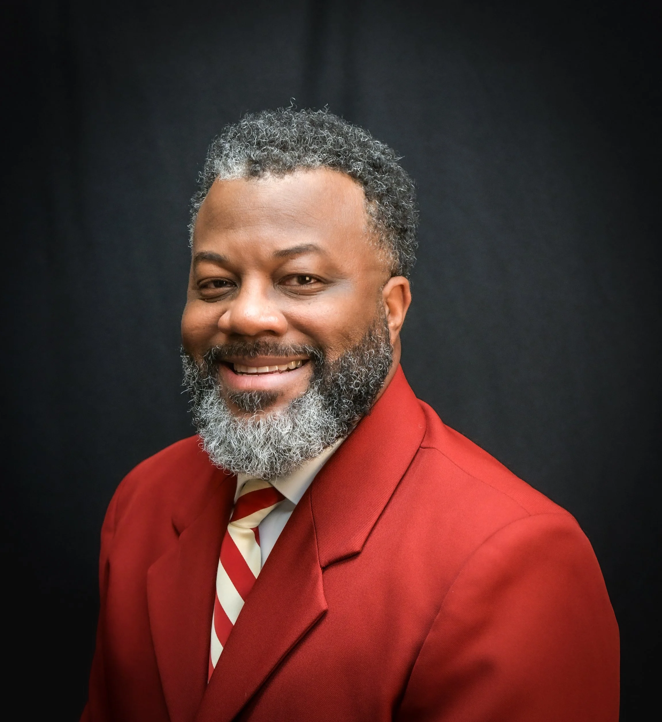 A man with a gray beard and short gray curly hair dressed in a red suit, white shirt, and red-and-white tie, smiling against a black background.