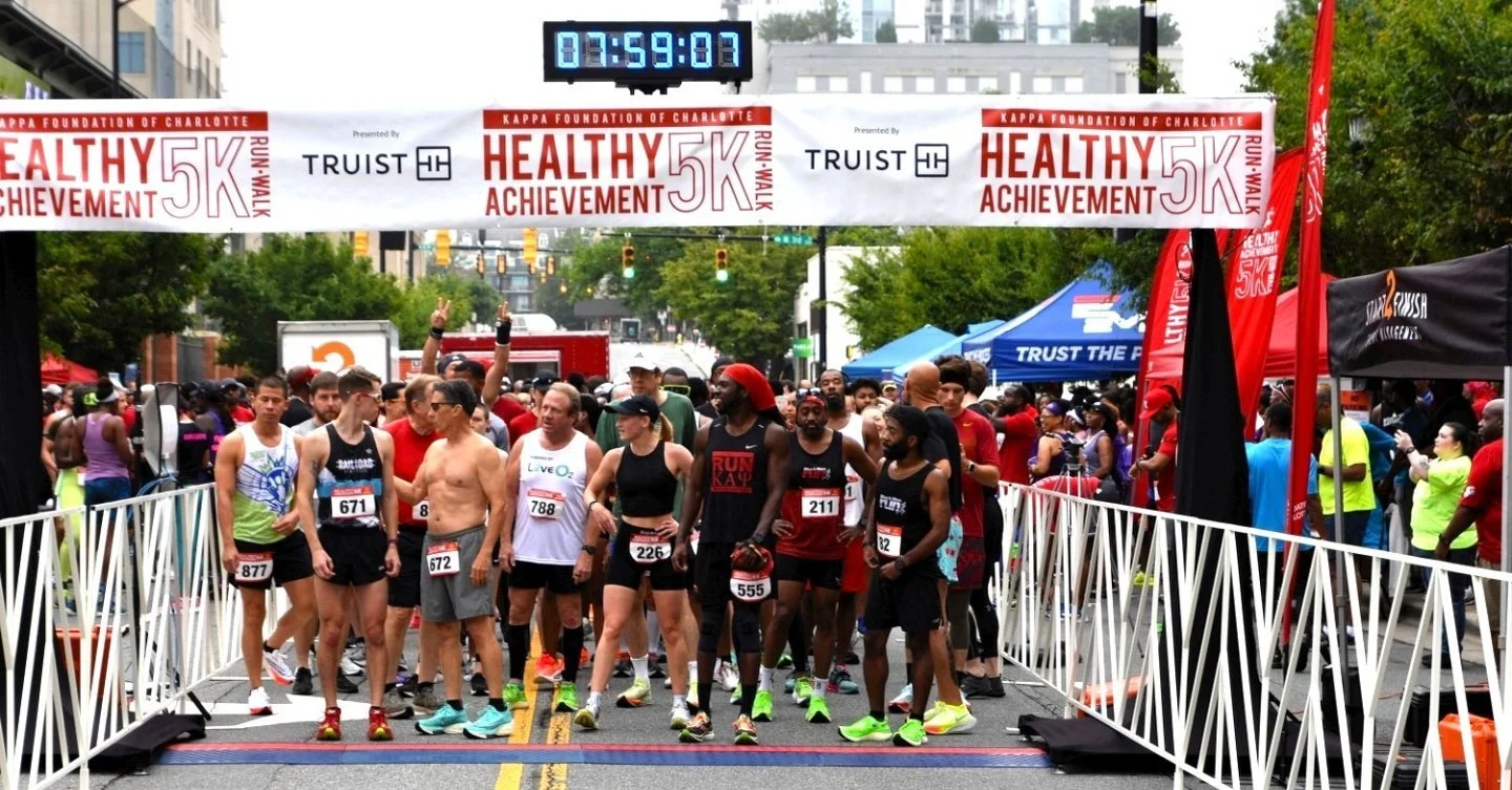 Start line of a 5K race with runners gathered behind a white barricade, banners overhead reading 'Healthy 5K' and 'Kappa Foundation of Charlotte', digital timer showing 59 minutes and 7 seconds, and crowd of spectators and event tents in the background.