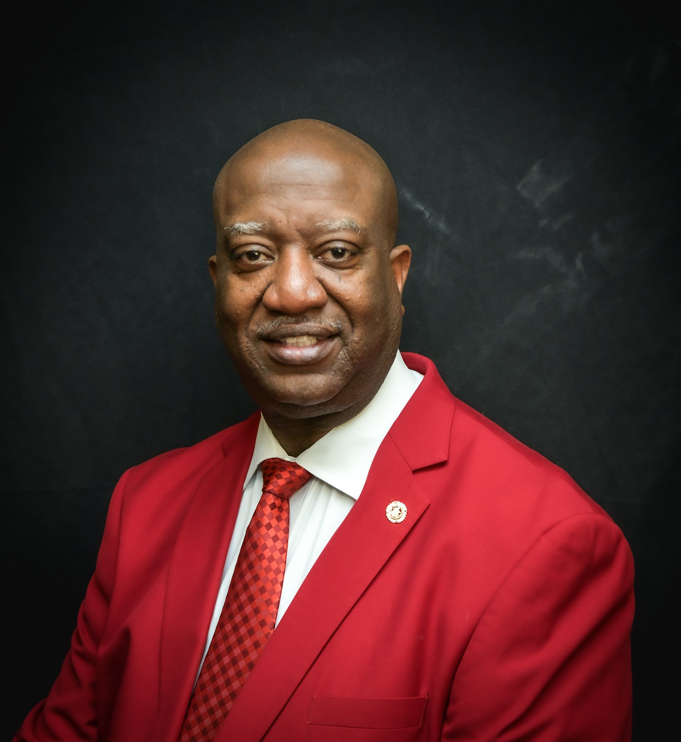 Portrait of an African American man wearing a red suit with a white shirt and a red checkered tie, standing against a dark background.