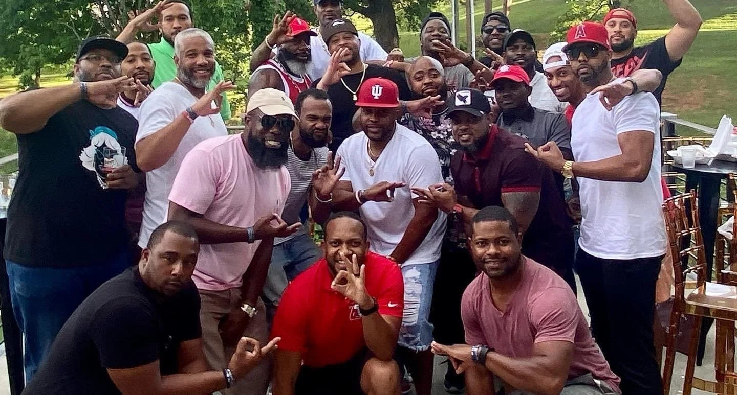 Group of men posing together outdoors; many are smiling and making hand gestures. Some wear caps, t-shirts, and sunglasses. The setting appears to be a park or patio with trees in the background.