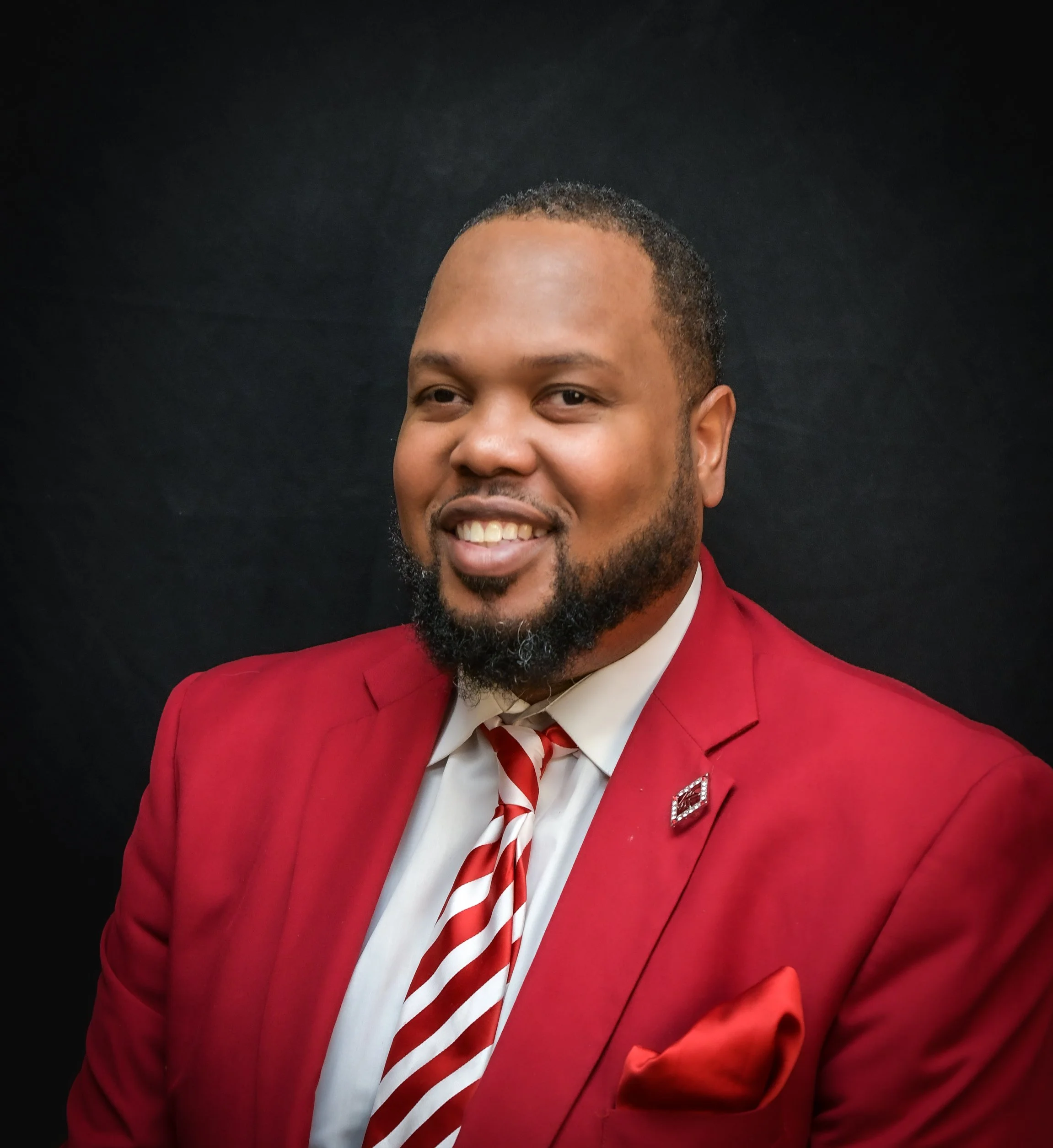 Close-up of a smiling man in a red suit and striped tie against a black background.