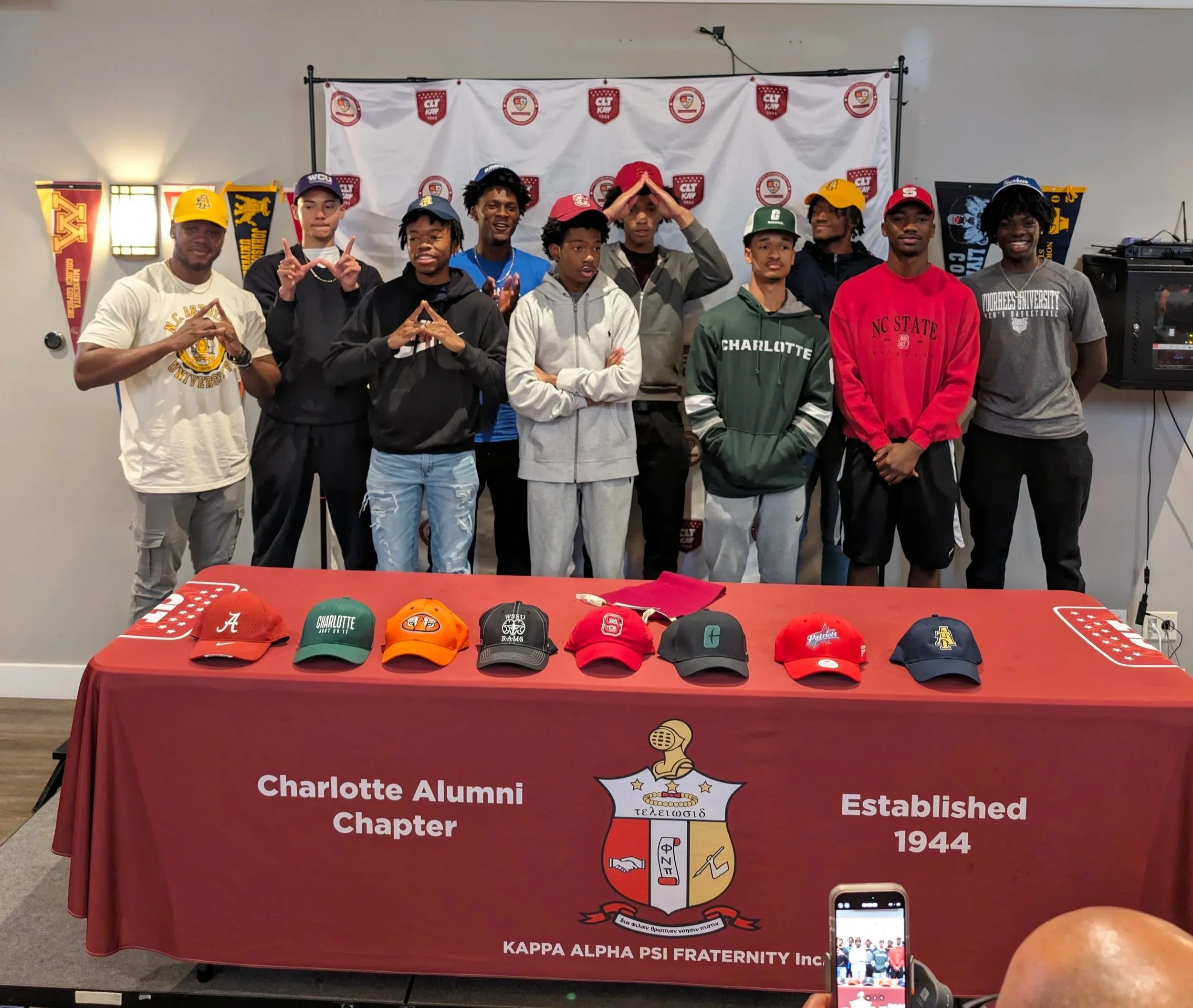 Group of young men standing behind a table with various caps on display, logo and banner of Charlotte Alumni Chapter of Kappa Alpha Psi Fraternity Inc., in a room with decorated walls.