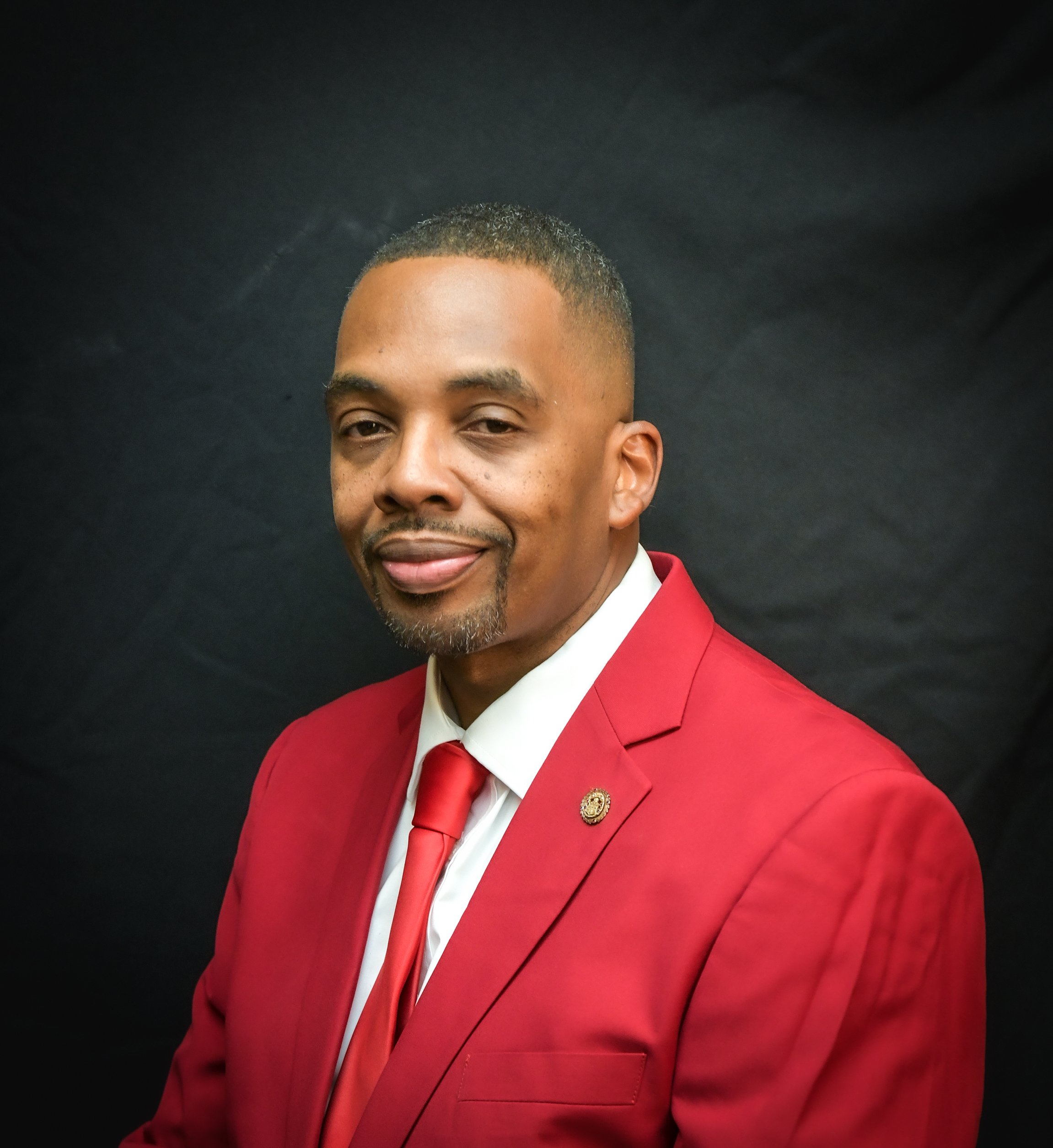 Close-up of a man wearing a red suit with a white shirt and red tie, smiling against a black background.