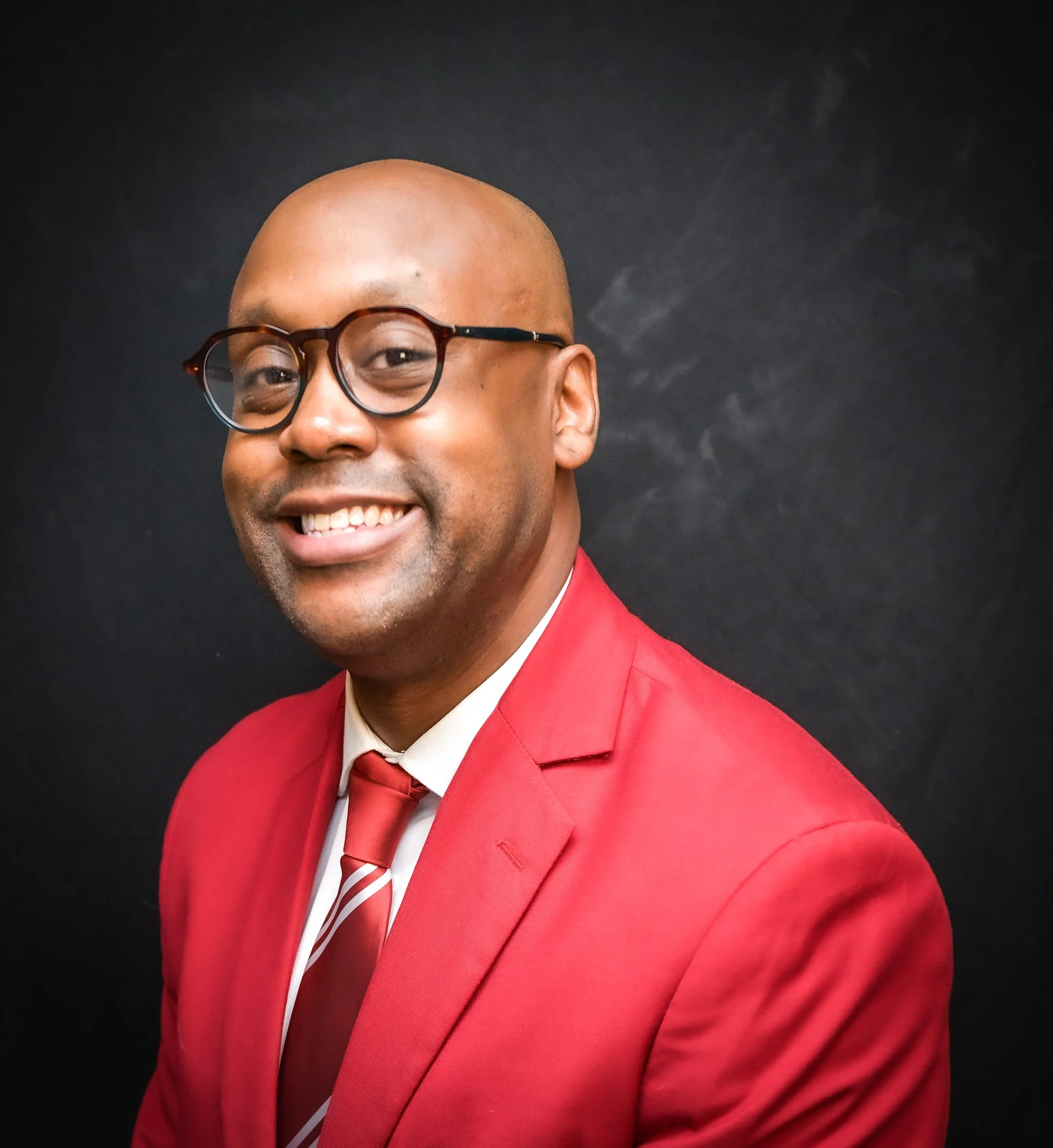 Close-up of a smiling Black man wearing round glasses, a red suit, and a matching red tie, against a dark background.