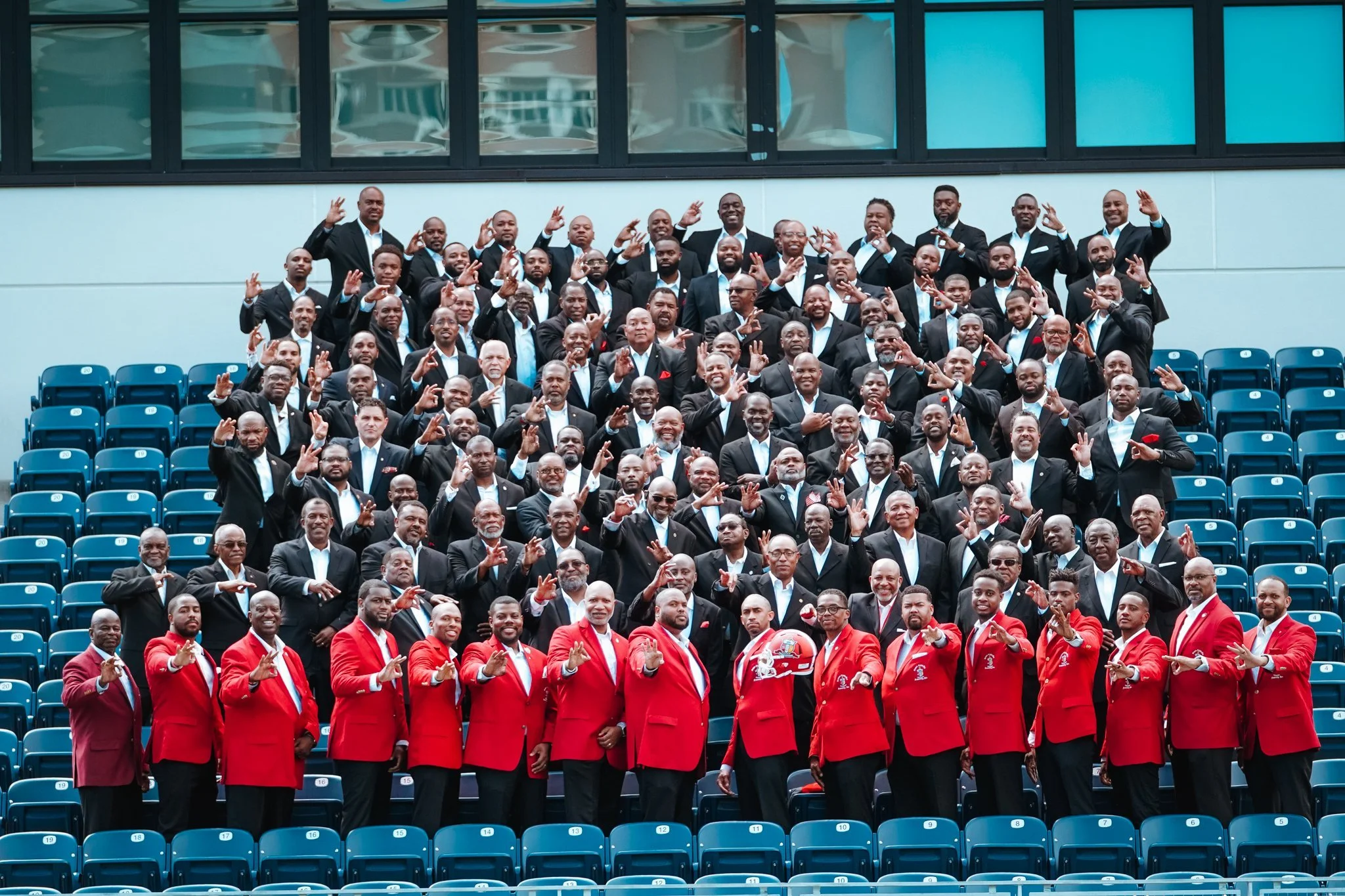 A large group of men dressed formally in suits and ties, some in red uniforms, posing for a group photo in an outdoor stadium with empty blue seats and a modern building in the background. Many men are making peace signs or gestures with their hands.