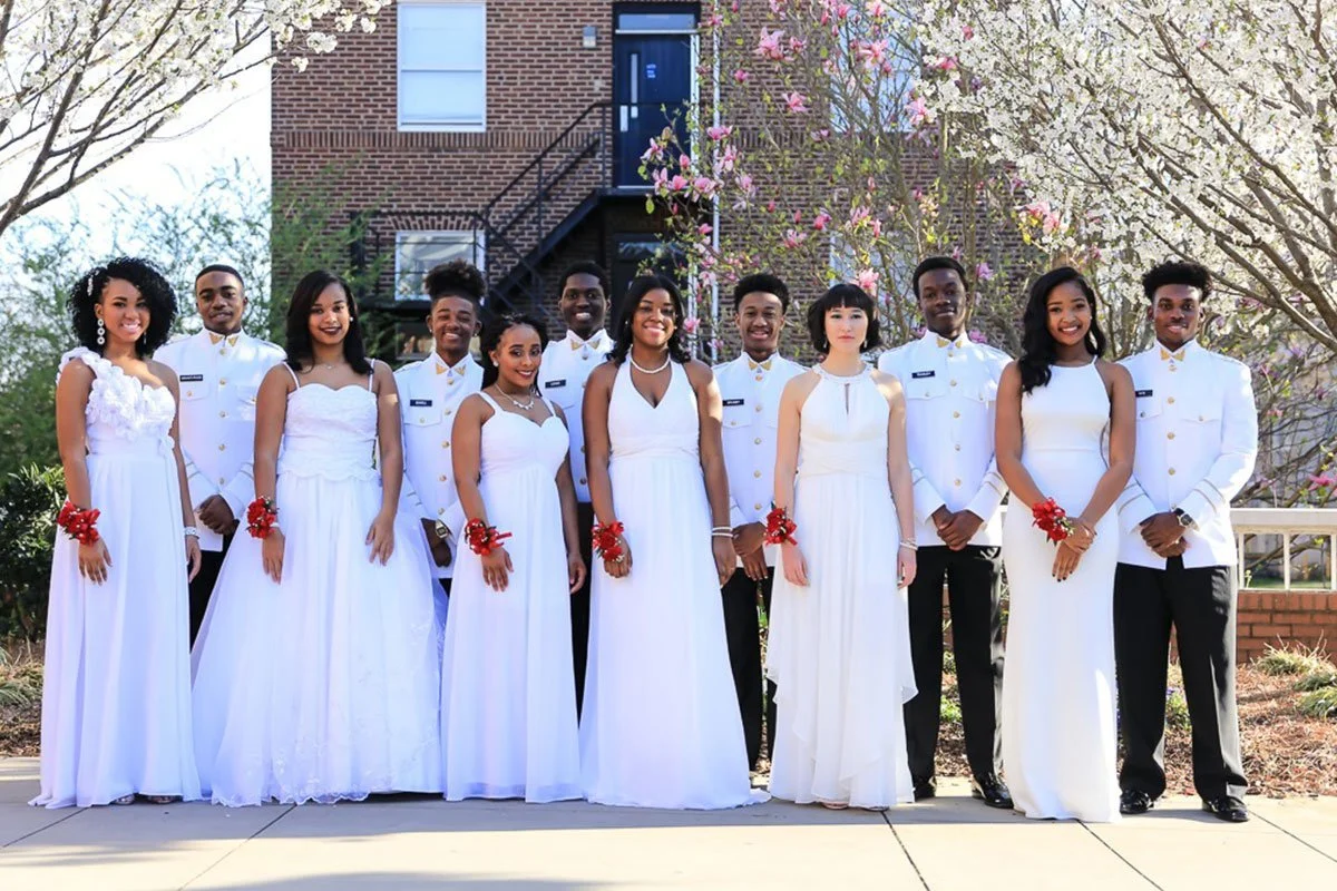 A group of people dressed in white formal attire standing outdoors in front of a brick building and flowering trees, smiling for a photo.