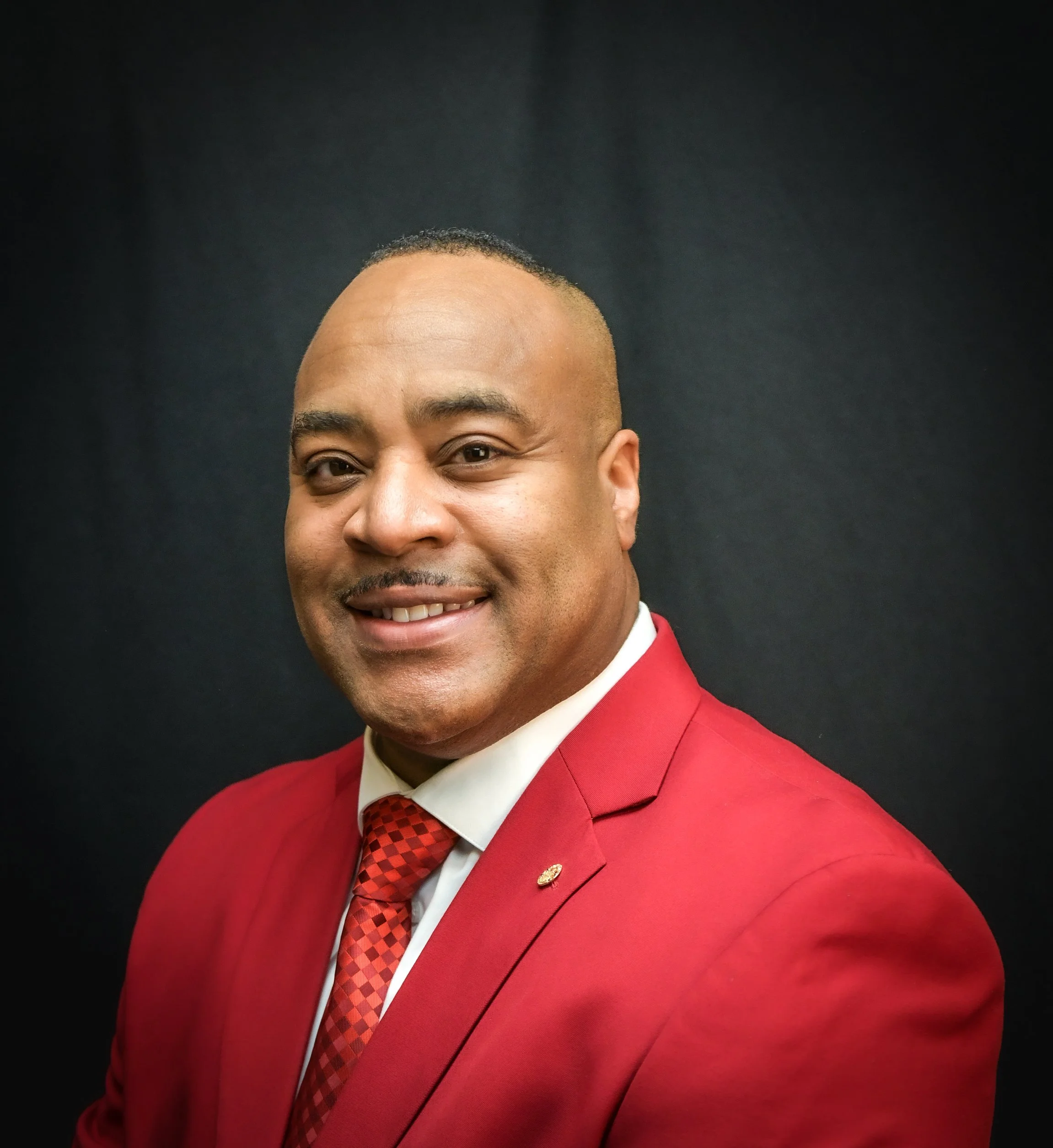 A man wearing a red suit, white shirt, red tie with black diamonds, and a red lapel pin, smiling against a dark background.