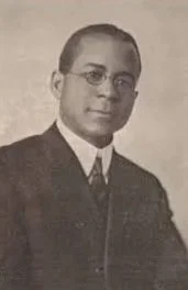 Black and white portrait of a young man wearing glasses, a dark suit, a white shirt, and a tie.