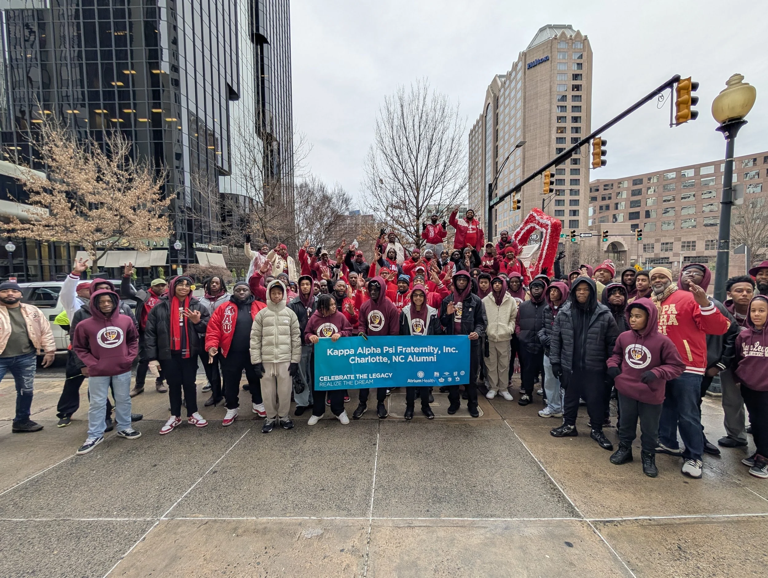 A diverse group of people gathered in a city street holding a blue banner that reads 'Kappa Alpha Psi Fraternity, Inc. Charlotte, NC Alumni' during a parade or event, with tall office buildings and traffic lights in the background.