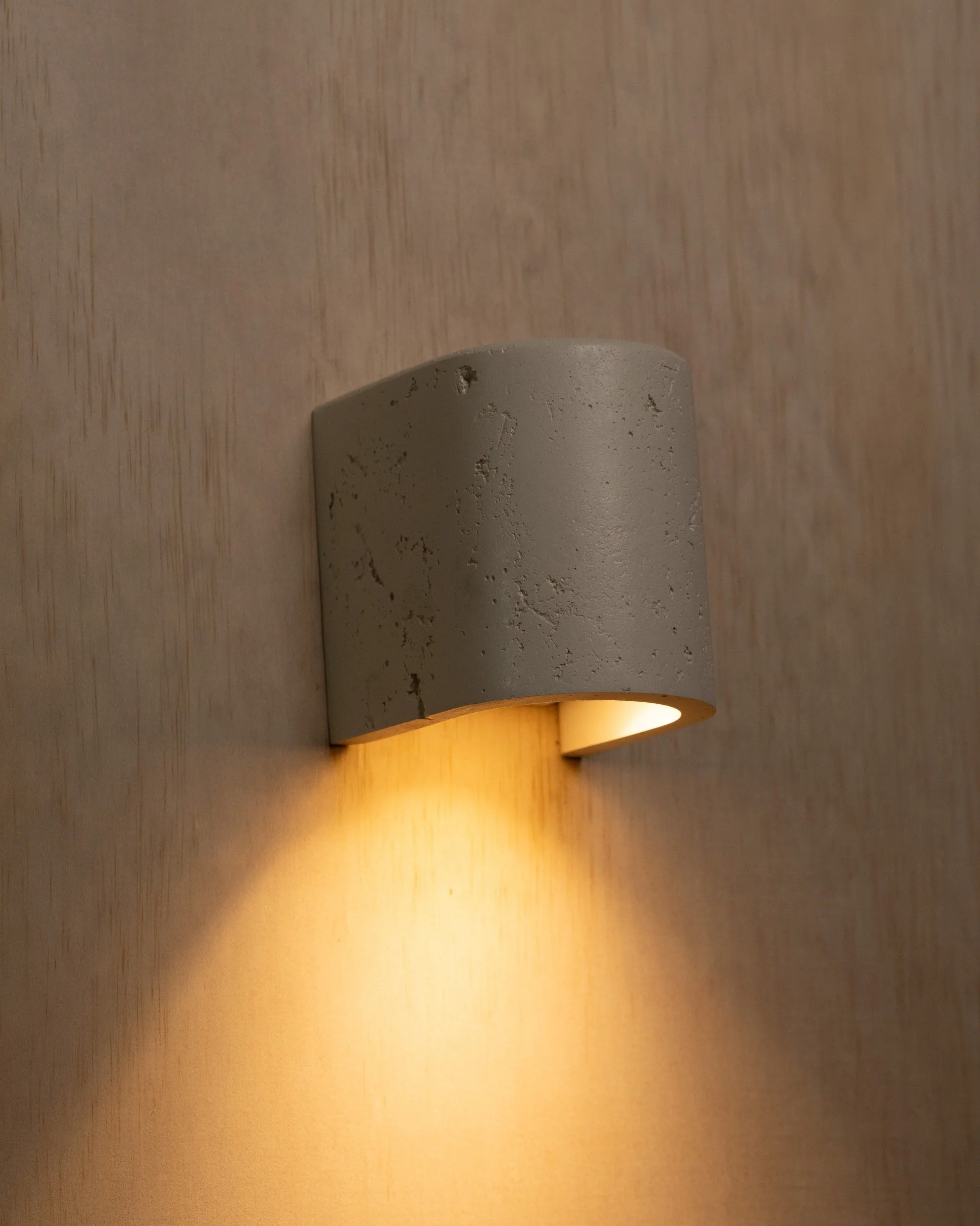 Close-up of a modern wall-mounted sconce with a cylindrical shape, textured surface, warm light illuminating downward, mounted on a light wood wall.