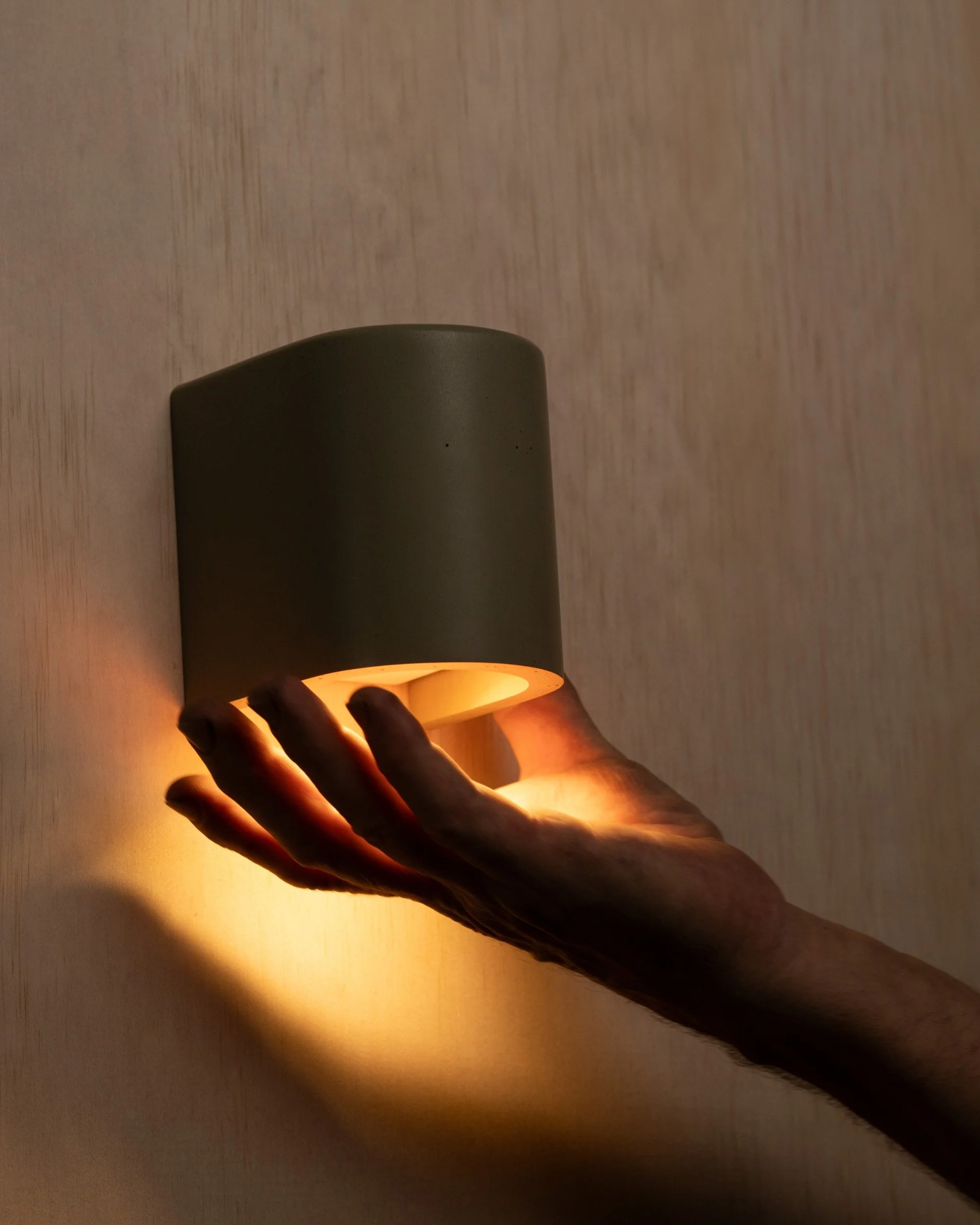 A person holding a cylindrical light fixture emitting warm light against a textured wall.