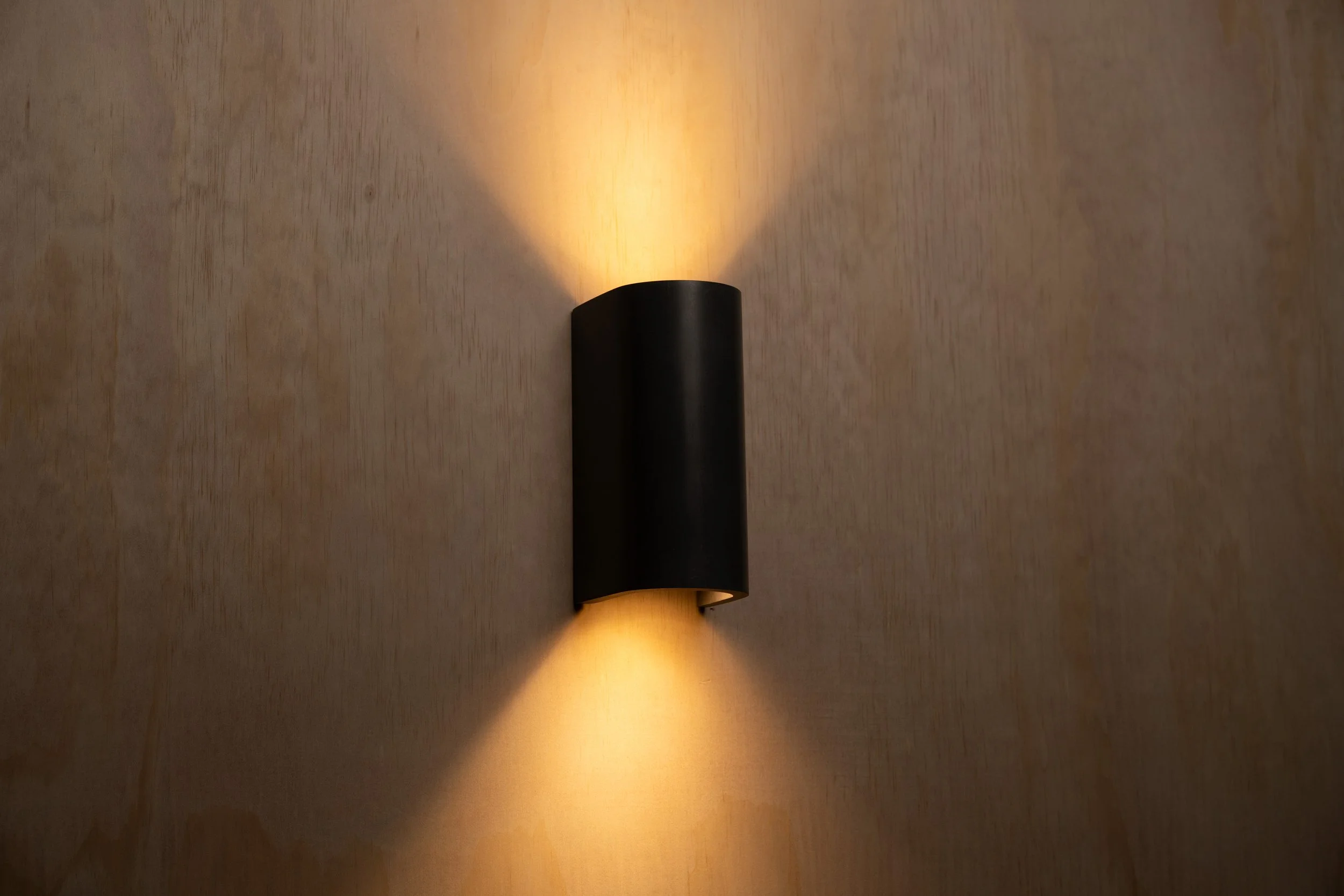 A modern wall-mounted light fixture with a black cylindrical body casting a warm, asymmetric glow on a textured wall.