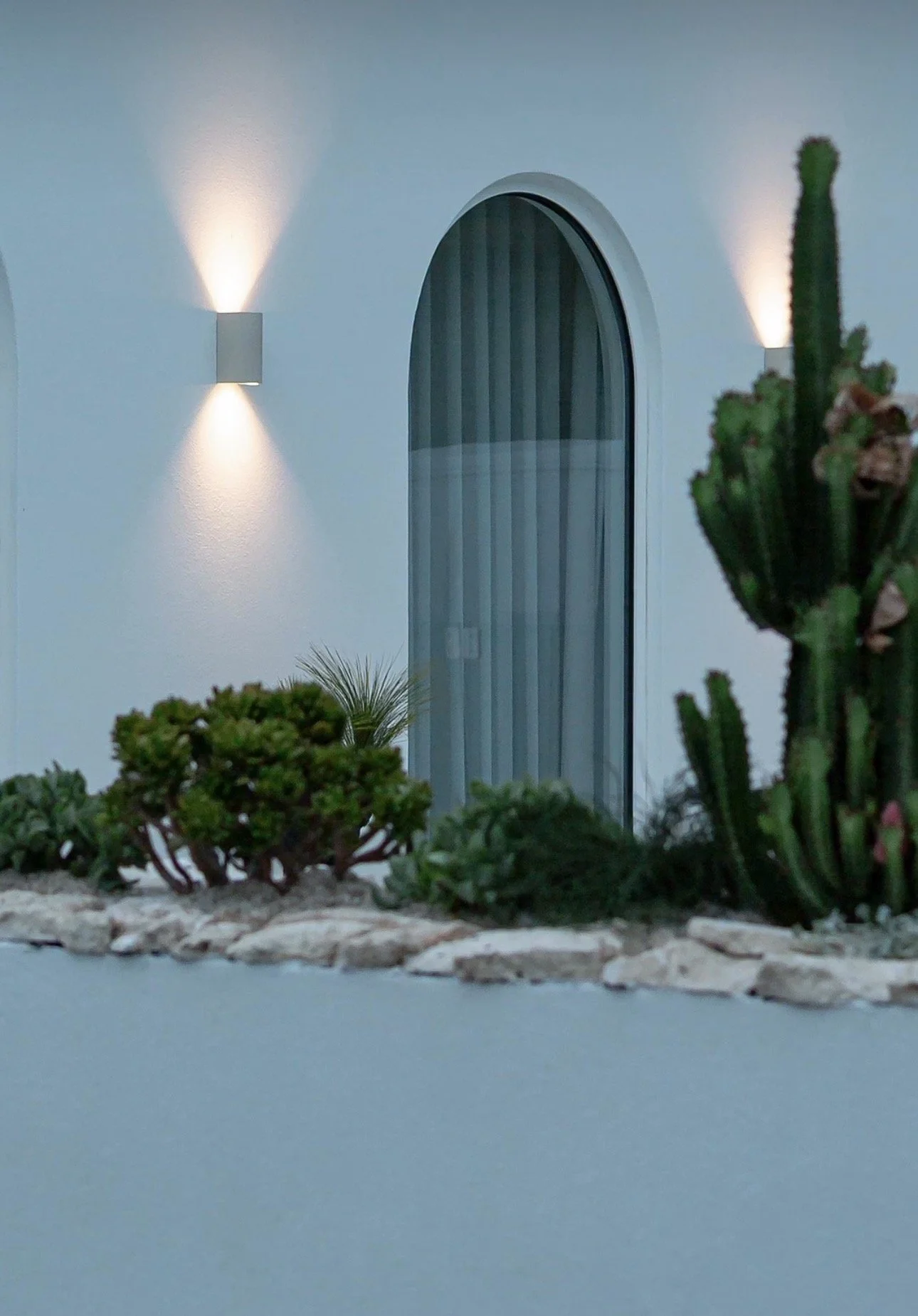 Exterior view of a modern house with white walls, a tall arched window with vertical blinds, and decorative wall-mounted lights. There are desert plants and succulents in a landscaped garden in the foreground.
