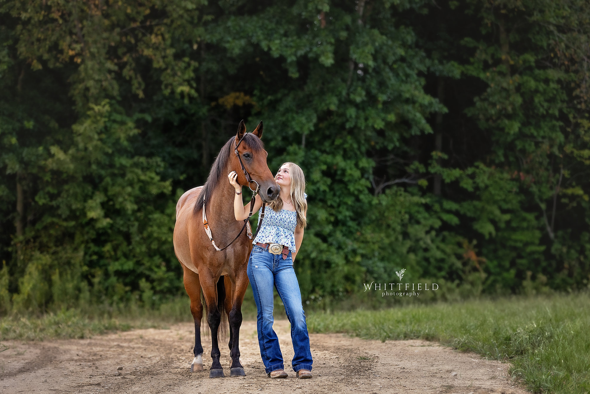 A woman with long blonde hair, wearing a blue and white floral blouse and jeans, standing on a dirt path next to a brown horse, with green trees in the background.