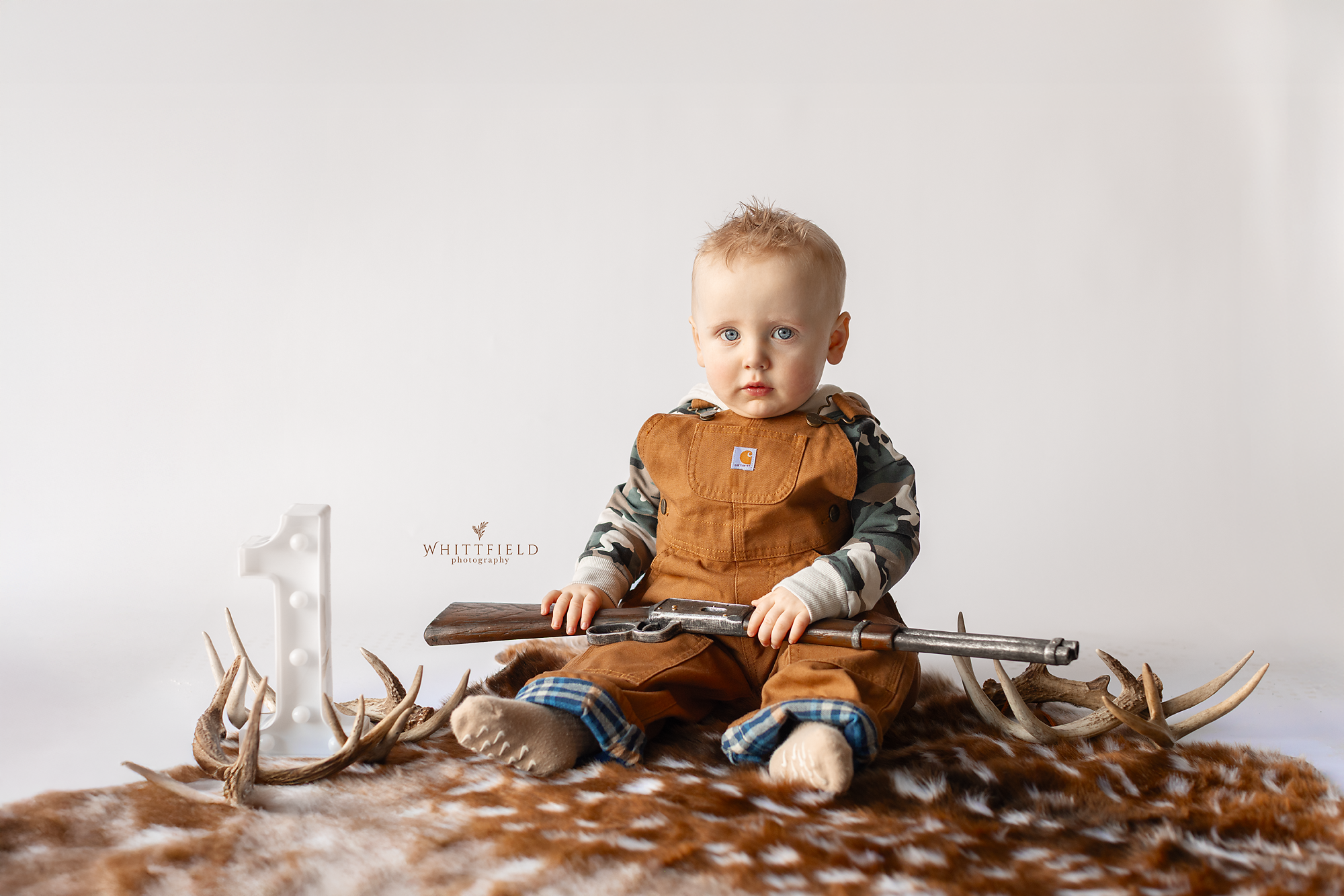 Young boy with blue eyes and light-colored hair sitting on a brown and white animal hide, holding a toy rifle, surrounded by deer antlers, with a white background and a small numeral '1' candle.