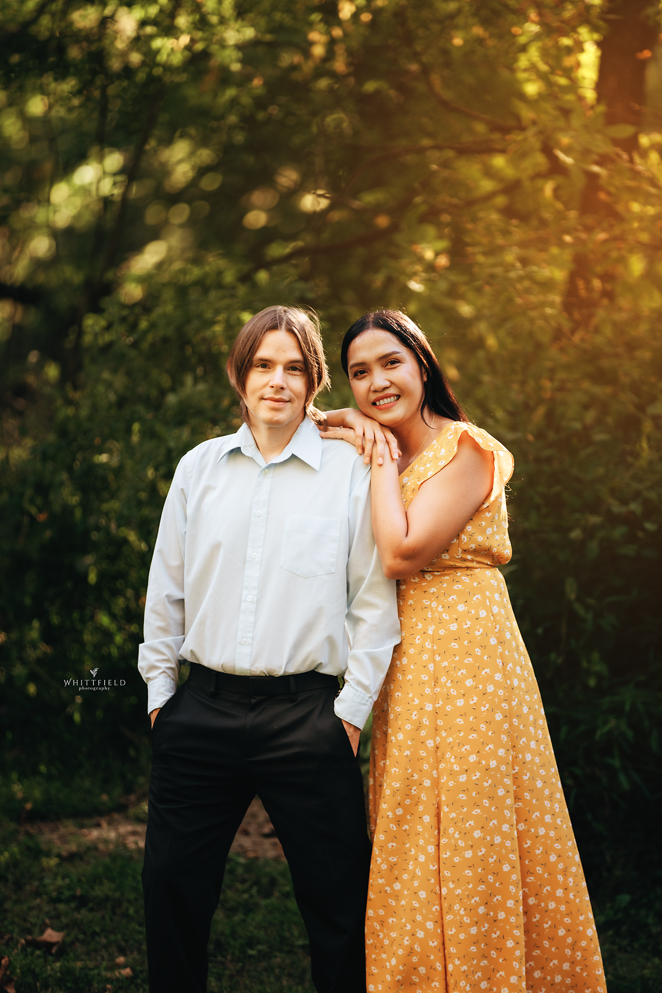 A young man and woman stand close together outdoors during sunset, with lush green trees in the background. The woman leans on the man’s shoulder, smiling, while the man looks calmly at the camera with hands in his pockets. The woman wears a yellow f