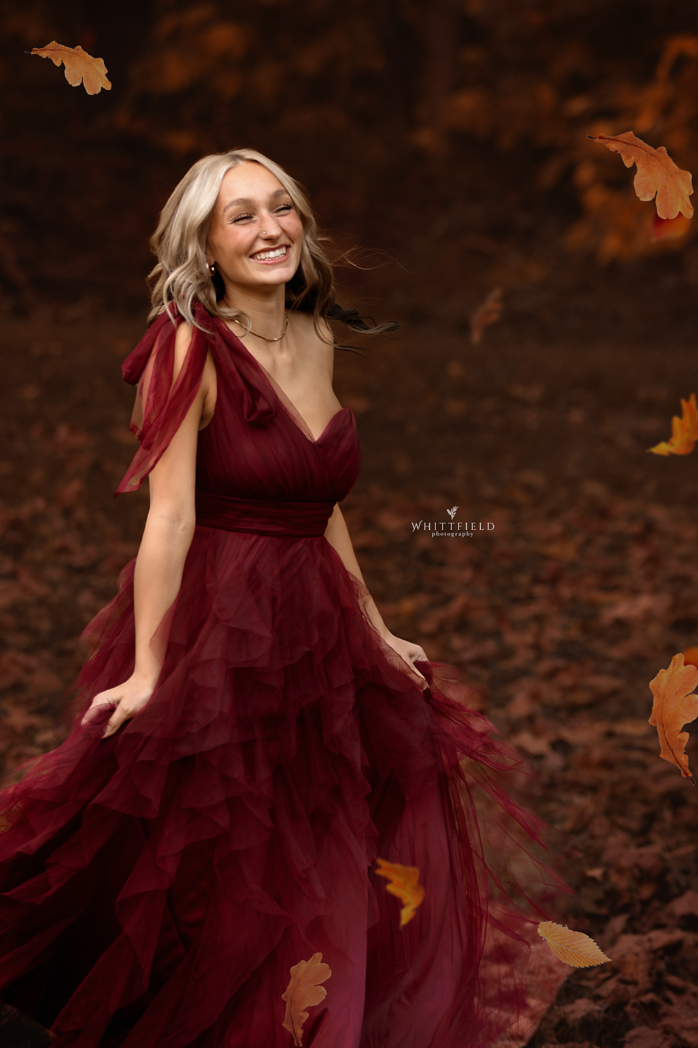 A woman in a flowing, ruffled red dress smiling in an autumn forest setting with falling leaves.