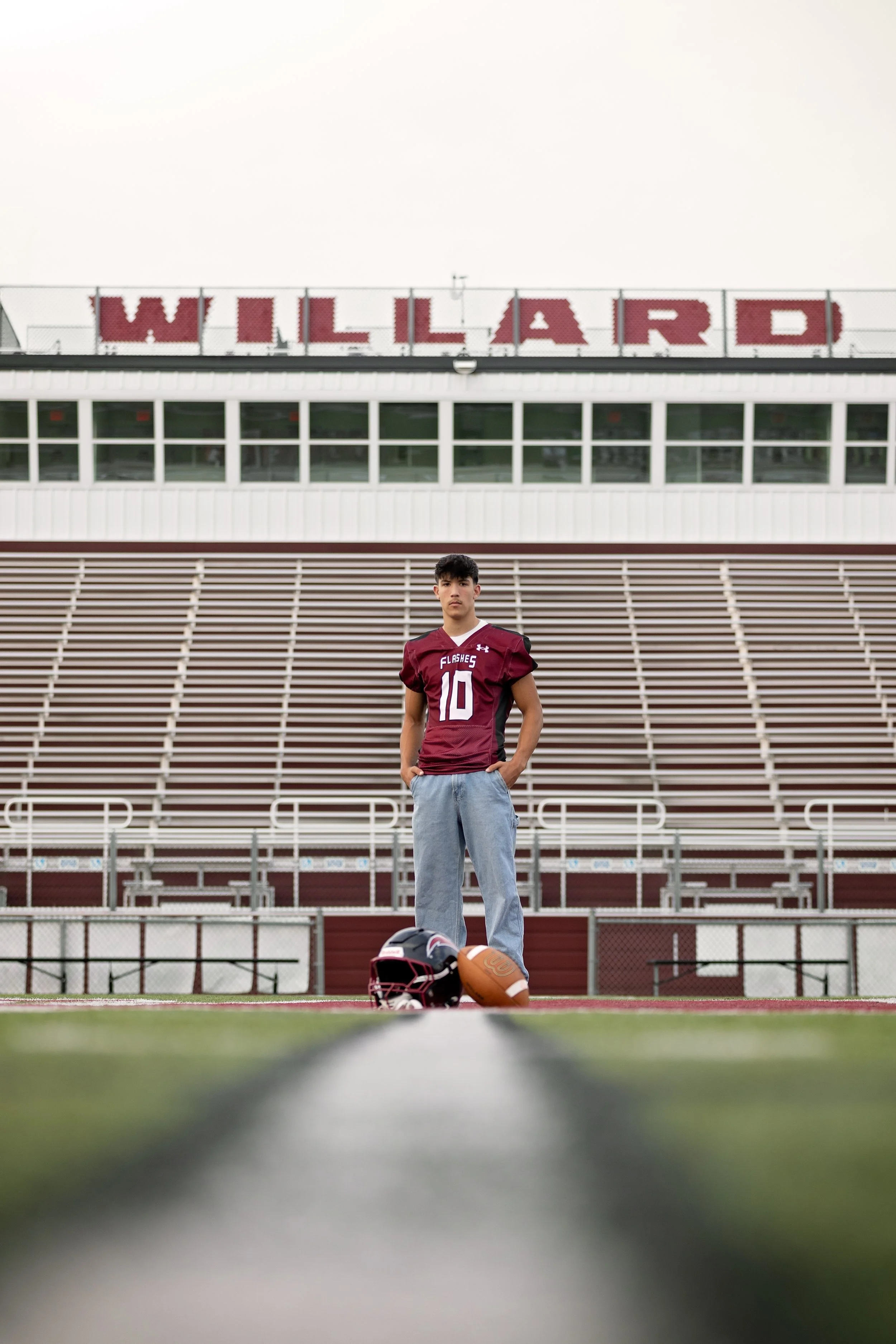 A young man standing on a football field wearing a maroon football jersey with the number 10, jeans, with a football helmet and football on the ground in front of him, stadium bleachers and a building with a large sign that reads 'WILLARD' in the bac