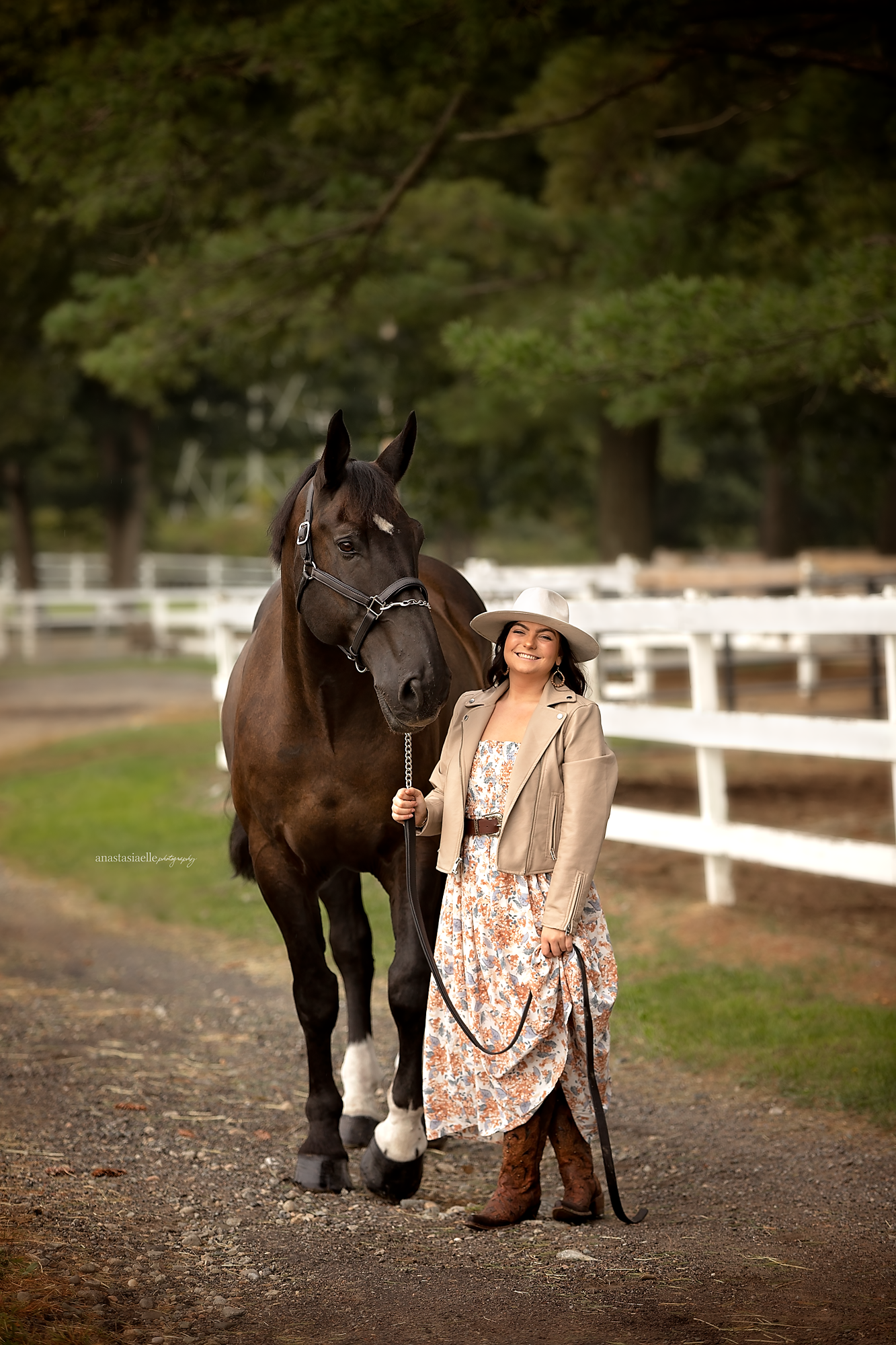 A woman in a floral dress, beige jacket, and cowboy boots stands beside a large dark brown horse with white markings on its feet and facial marking, holding its reins, in a rural setting with white fences and tall green trees in the background.