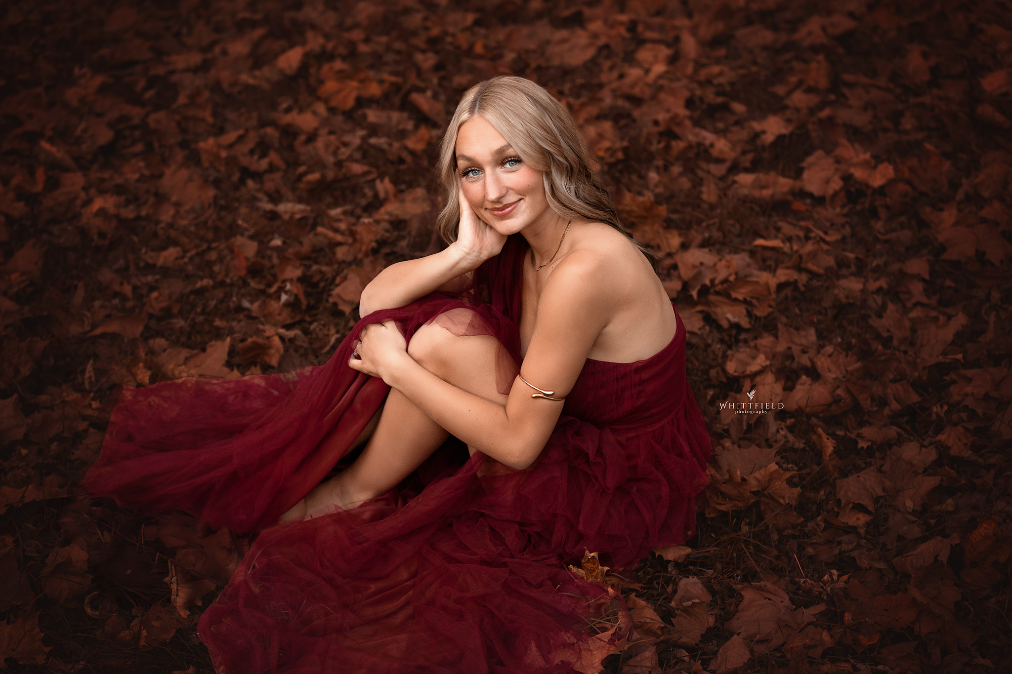 A young woman with blonde hair is sitting on the ground covered with brown autumn leaves, wearing a strapless red dress, and smiling at the camera.