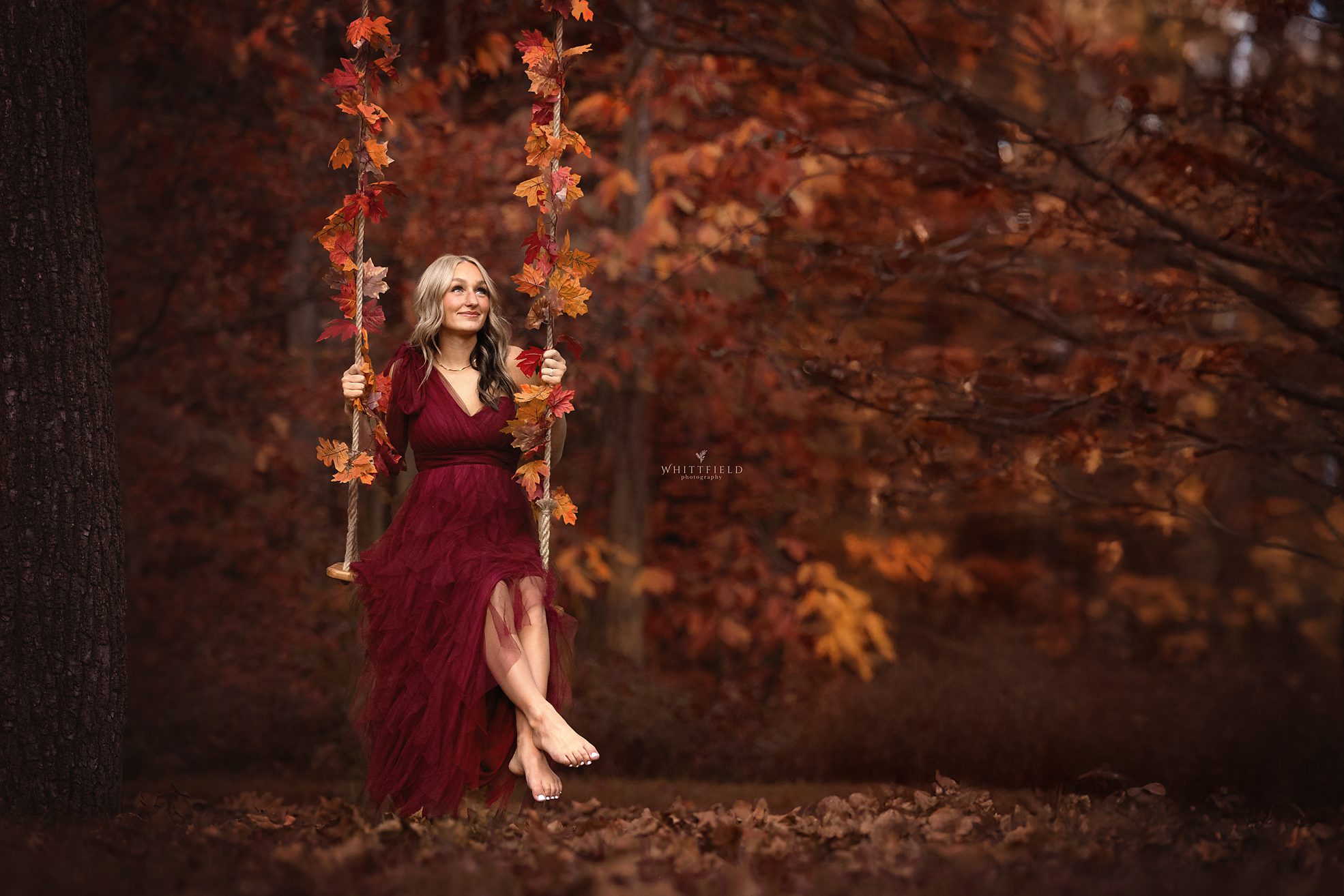 A woman in a long, burgundy dress sitting on a swing decorated with autumn leaves, in a forest with orange and red fall foliage.