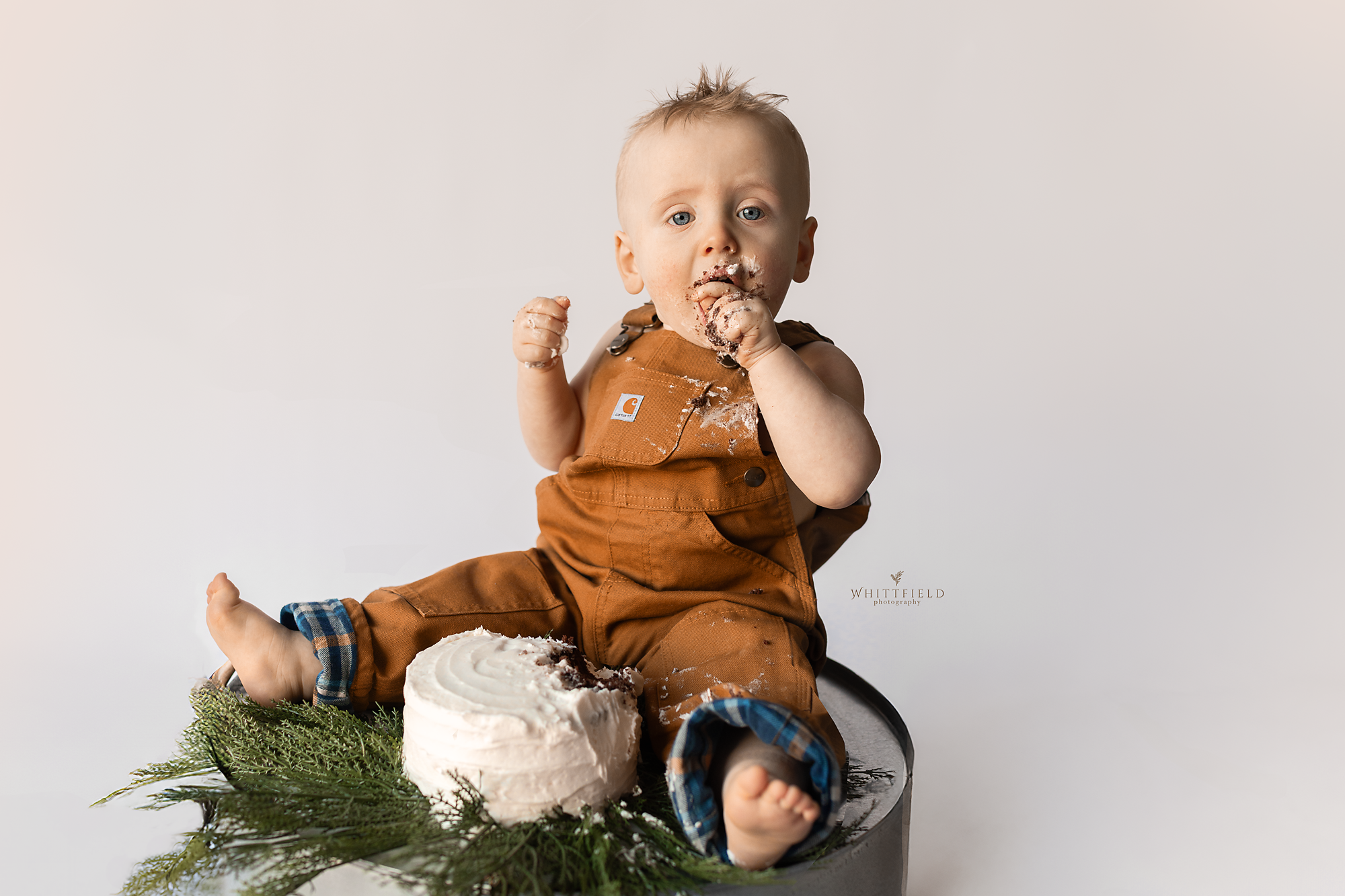 A young child with messy face and hands sitting on a surface with a small cake and greenery, wearing brown overalls, looking at the camera.